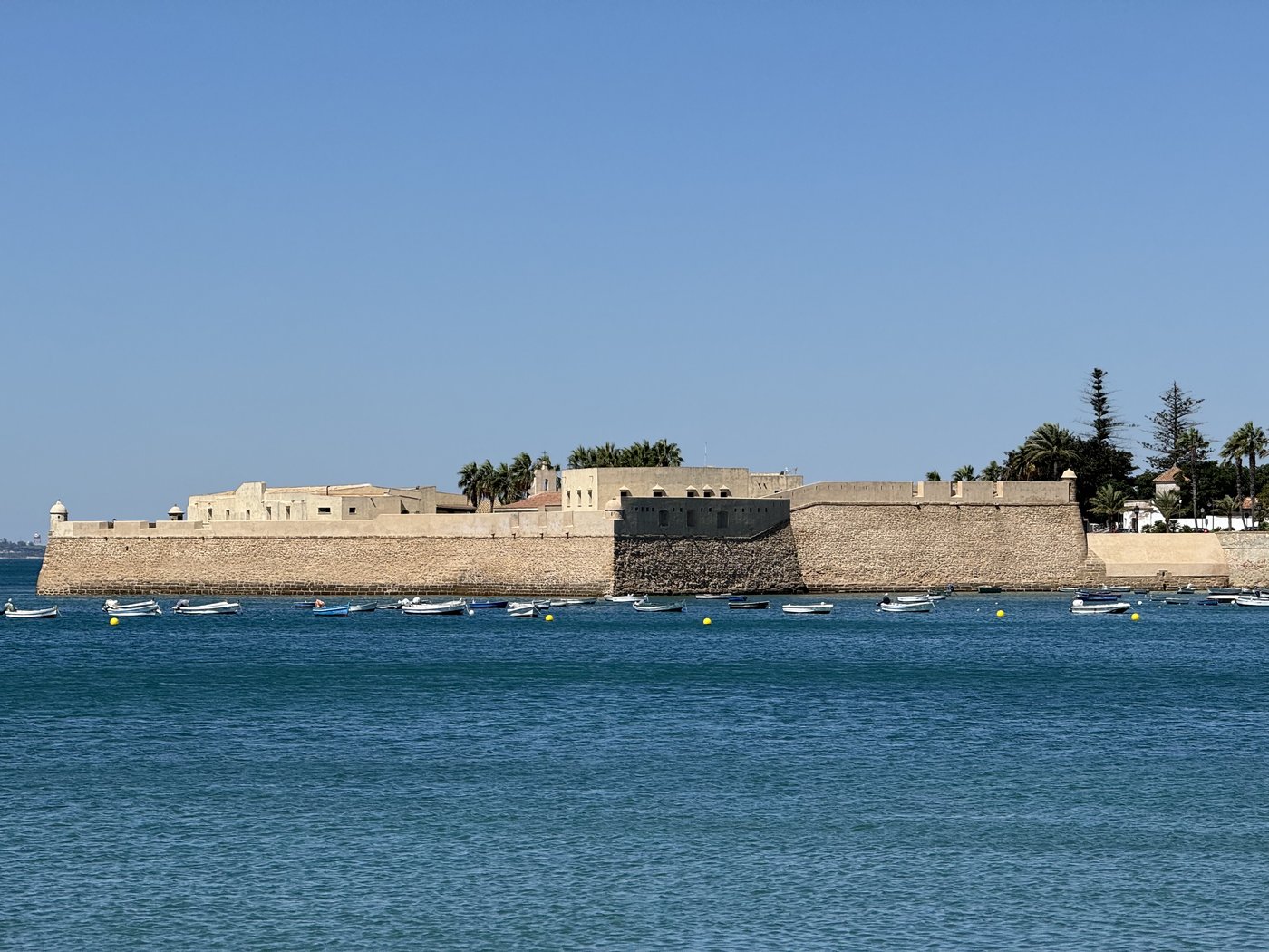 Das Castillo de San Sebastián auf seiner Felseninsel: niedrige Festungsmauern aus hellem Stein, ringsum türkisfarbenes Wasser unter wolkenlosem Himmel.