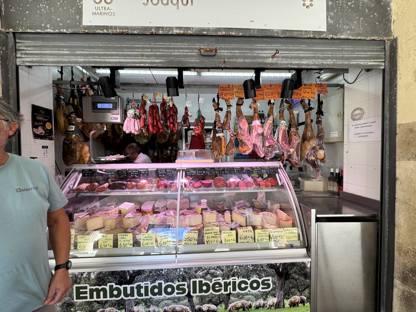 Eine Kühlvitrine voller Wurst- und Schinkenwaren mit der Aufschrift Embutidos Ibéricos, darüber hängen ganze luftgetrocknete Schinken von der Decke.