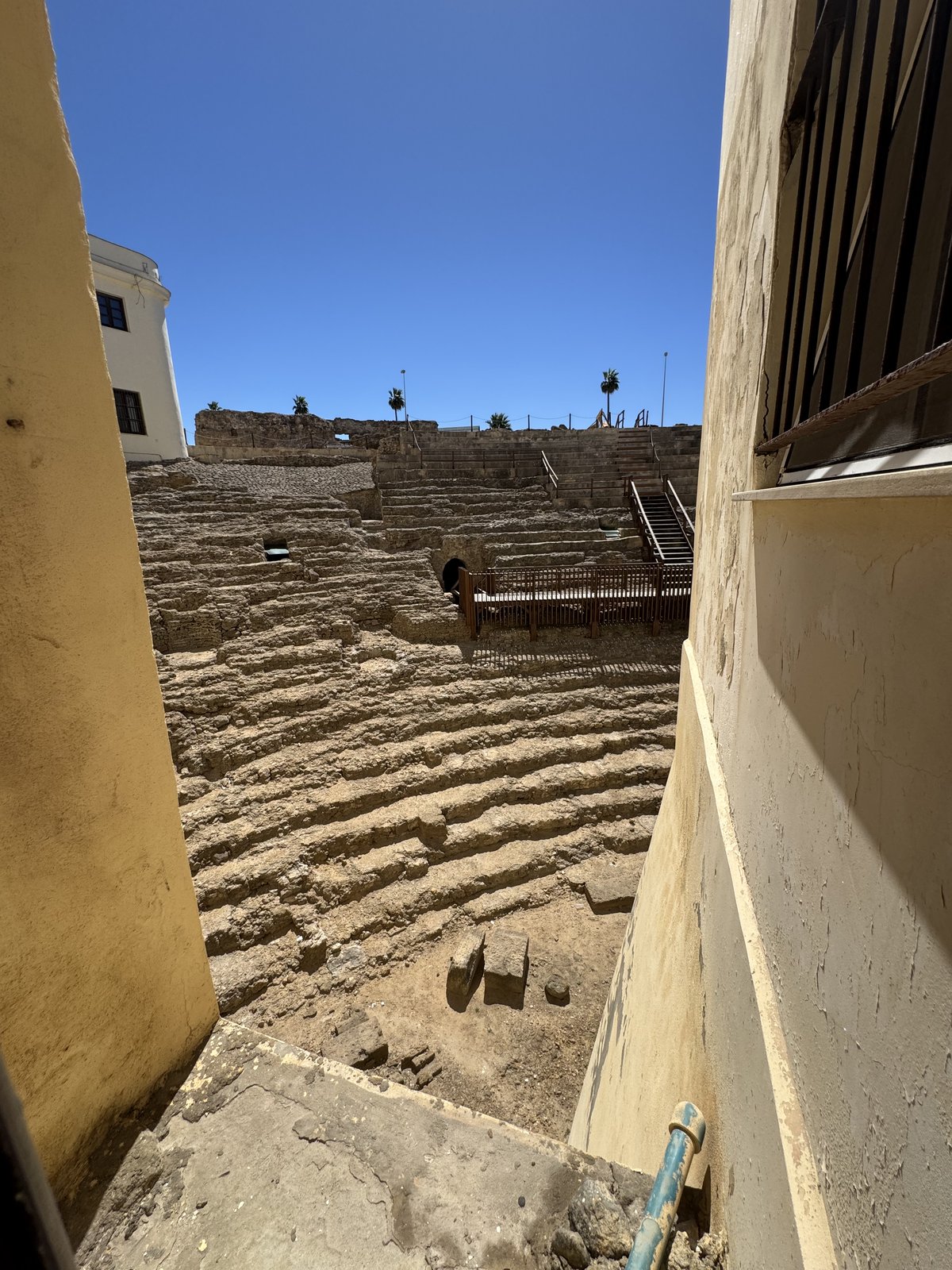 Durch eine Maueröffnung hindurch der Blick auf die freigelegten steinernen Sitzreihen des römischen Theaters, hell in der Mittagssonne.
