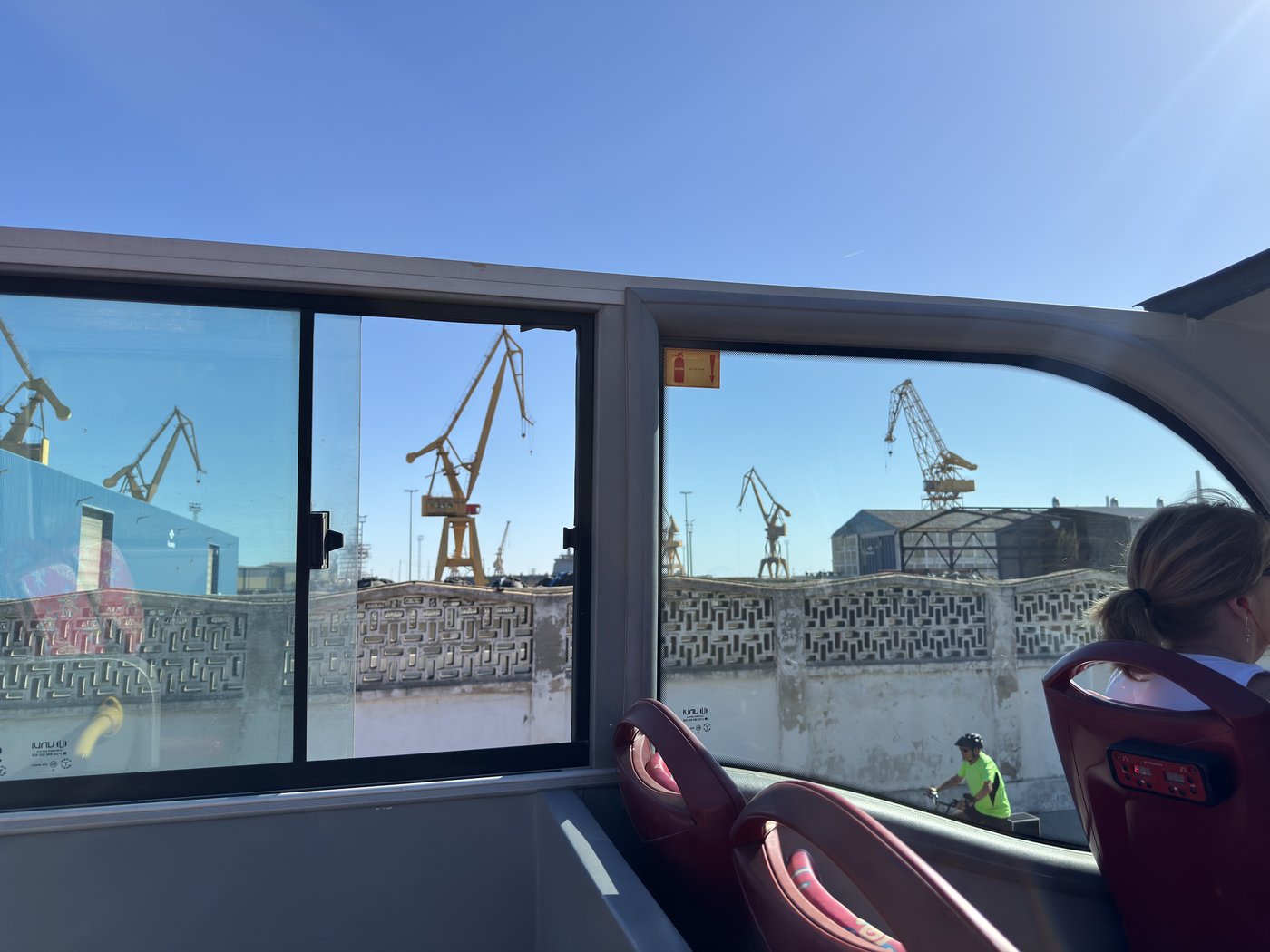 Blick aus dem Fenster des offenen Sightseeing-Busses auf das Hafengelände mit Containerkränen, die Sitzreihen davor sind fast leer.