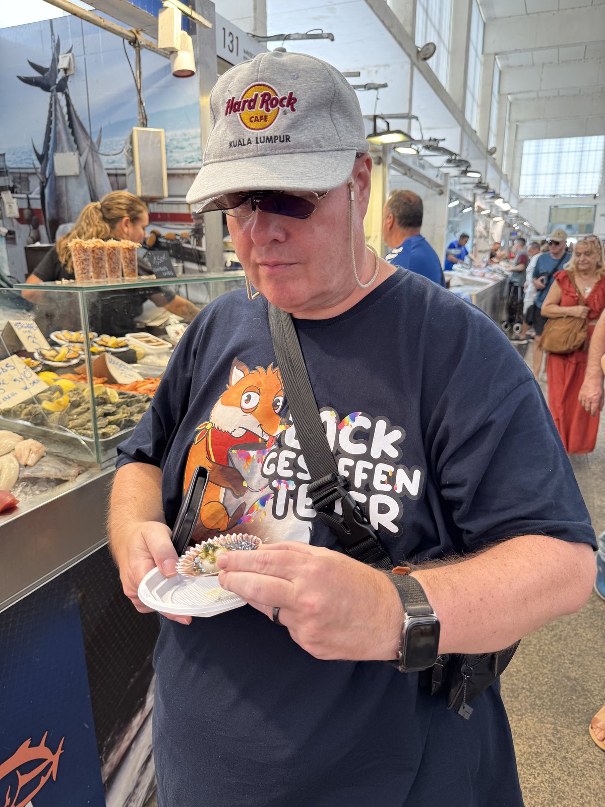Henning mit heller Kappe hält in der Markthalle ein kleines Pappschälchen mit einer Muschel in der Hand und betrachtet sie skeptisch.