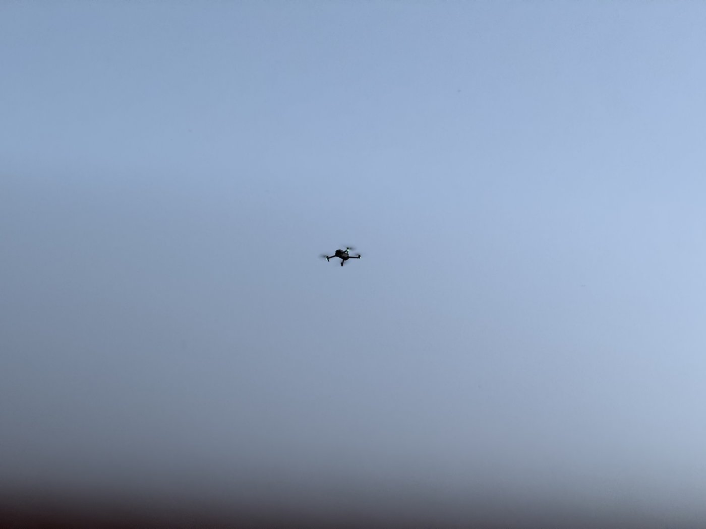 Eine kleine Drohne mit vier Rotoren schwebt als dunkle Silhouette vor einem dämmrigen, wolkenlosen Himmel.