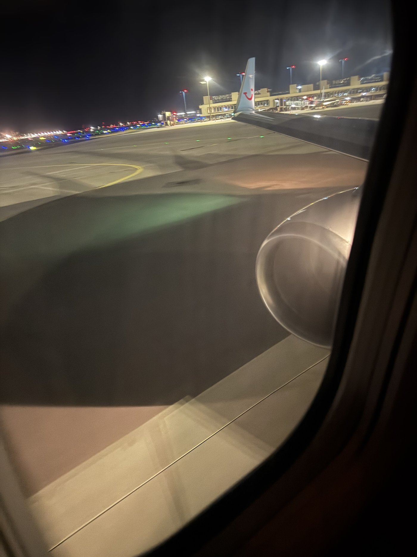 Blick aus einem Flugzeugfenster auf ein nächtliches Vorfeld: Lichterketten, Fahrzeuge und Gebäude in der Dämmerung.