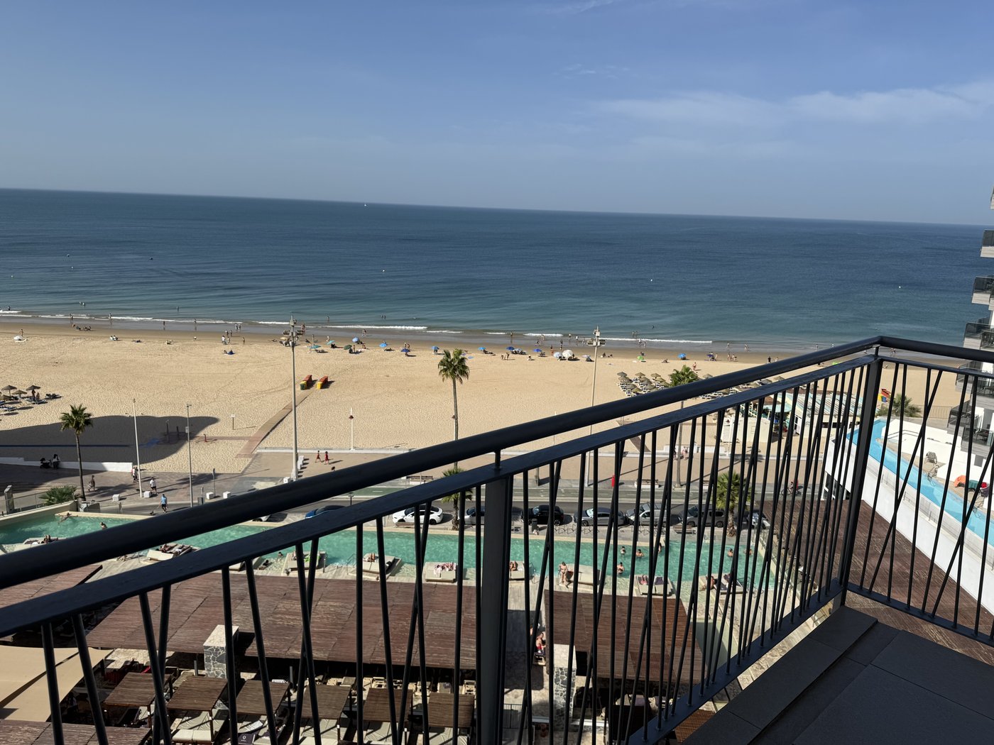Blick von einem Hotelbalkon über einen Pool auf einen breiten Sandstrand und das Meer unter blauem Himmel.