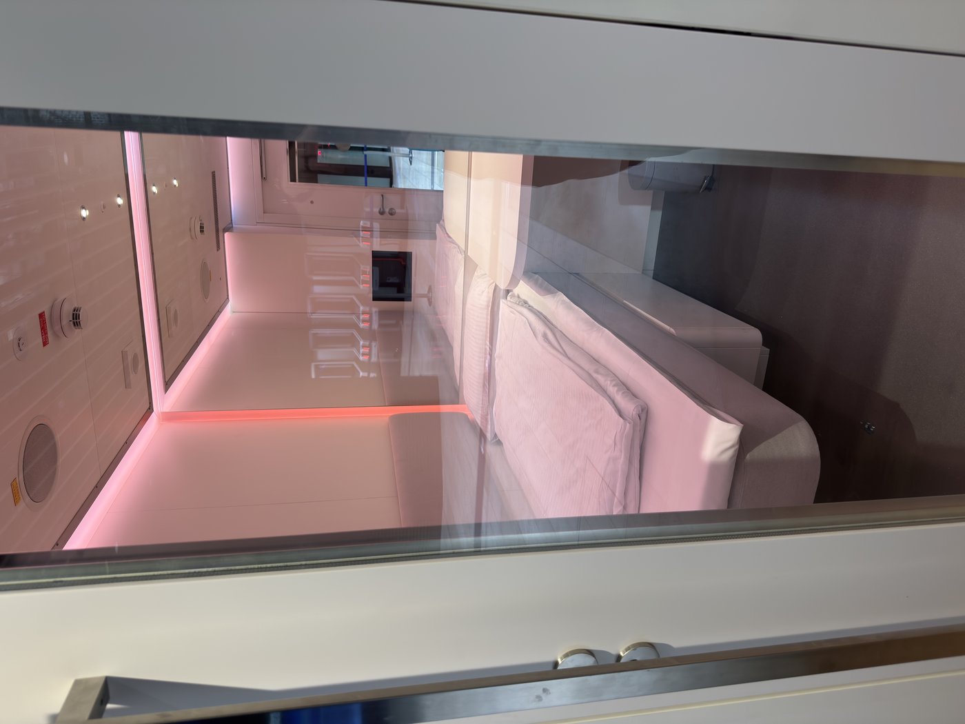 Eine winzige Schlafkabine hinter einer Glasscheibe: ein schmales Bett unter rosafarbenem Licht, an der Wand ein Bildschirm.