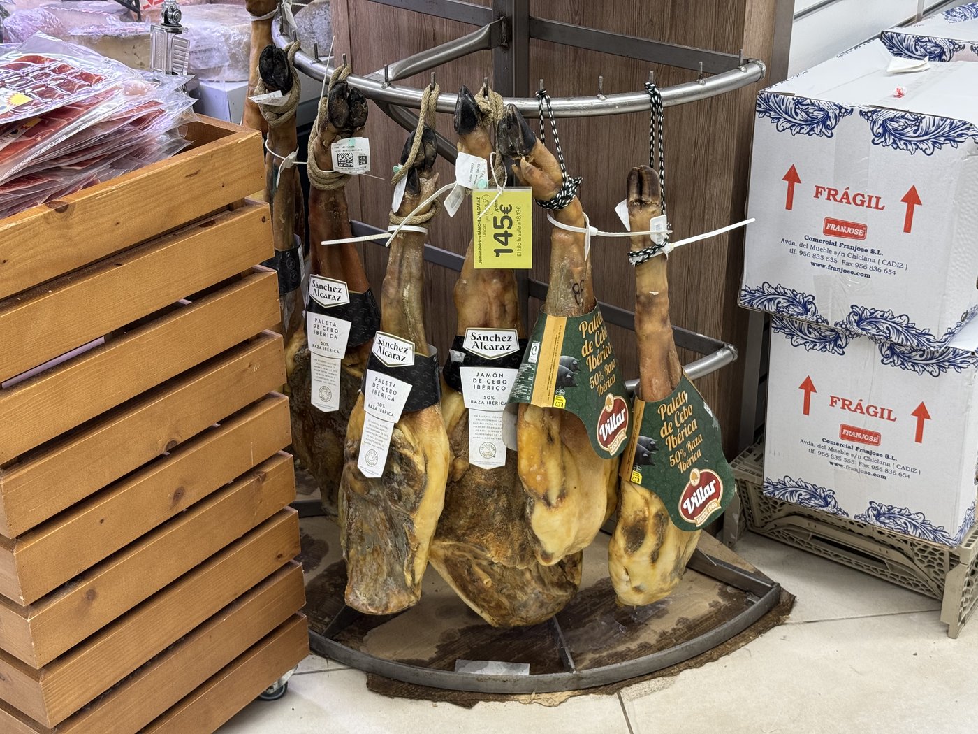 Mehrere ganze luftgetrocknete Schinken hängen an einem hölzernen Ständer in einem Supermarkt, daneben Regale mit Waren.
