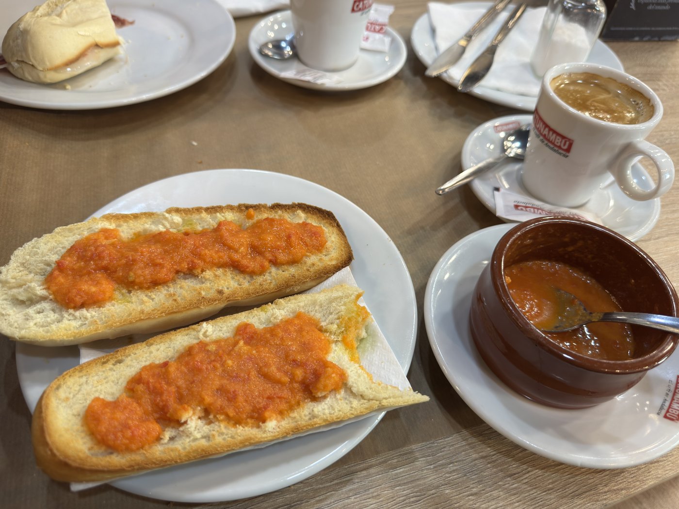 Zwei aufgeschnittene geröstete Brote mit zerdrückten Tomaten auf einem Teller, daneben eine Tasse Milchkaffee und ein Cortado.
