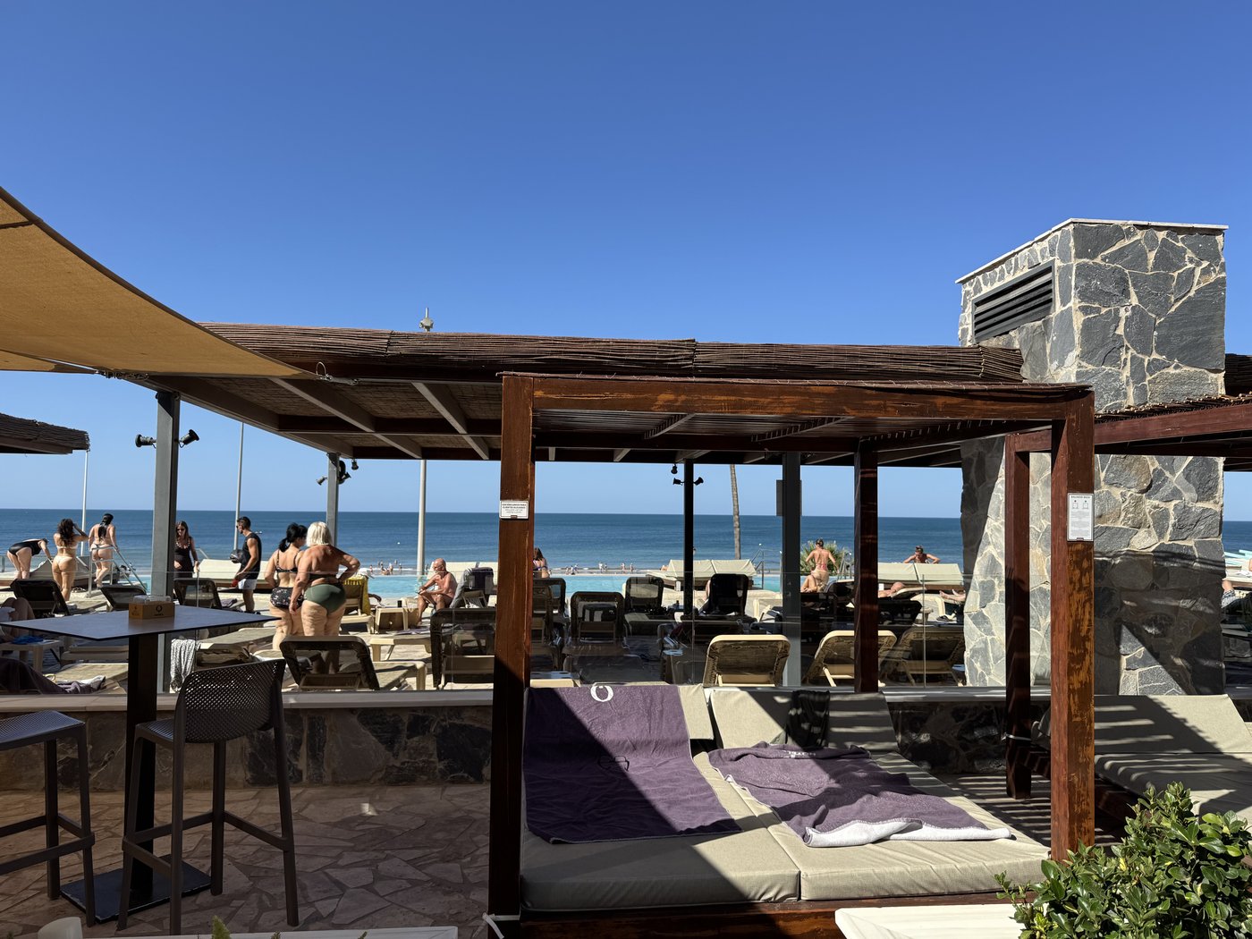Eine Hotelterrasse mit Sonnenliegen unter hellen Sonnensegeln, im Hintergrund das Meer unter wolkenlosem Himmel.