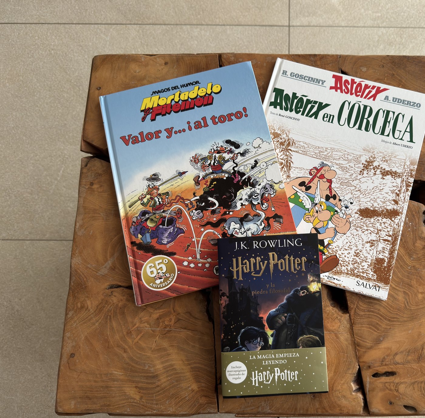 Drei Bücher auf einem rustikalen Holztisch, von oben gesehen: ein Comicband von Mortadelo y Filemón, Astérix en Córcega und ein Harry-Potter-Band auf Spanisch.