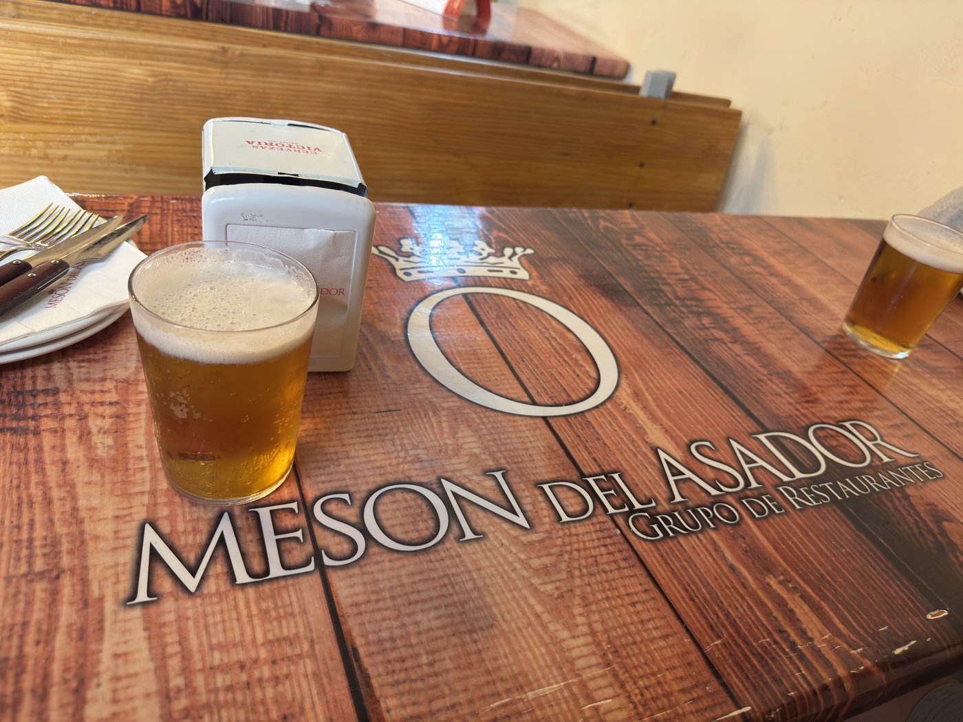 Ein Holztisch mit eingebranntem Logo Mesón del Asador, darauf ein Glas helles Bier und eine Serviettenbox.