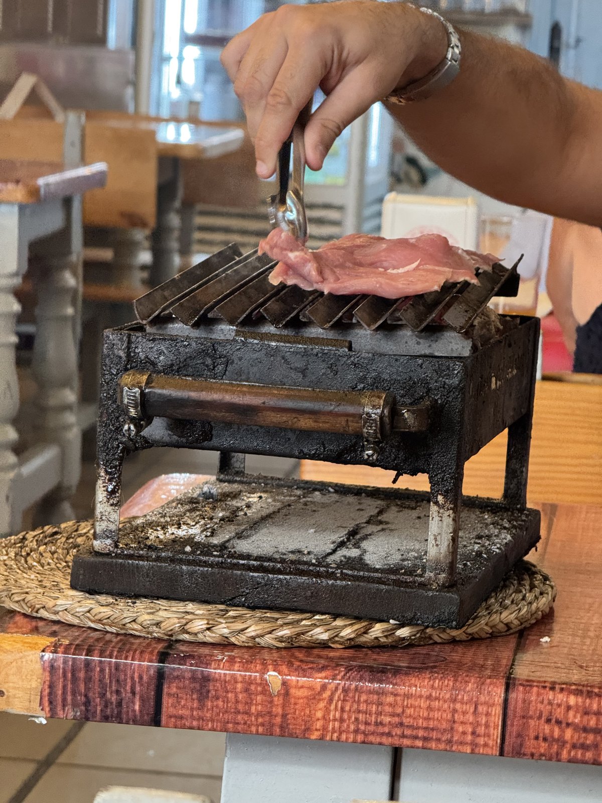 Ein kleiner gußeiserner Holzkohle-Tischgrill auf einem Strohuntersetzer; eine Hand wendet mit einer Gabel ein Stück Fleisch darauf.