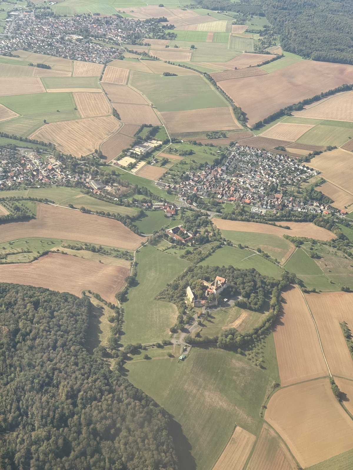 Blick aus dem Flugzeug kurz vor der Landung: ein Flickenteppich aus grünen und abgeernteten Feldern, Waldstücke und Dörfer. Unten auf einem bewaldeten Hügel liegt eine Burganlage mit hellen Mauern und rotem Dach.