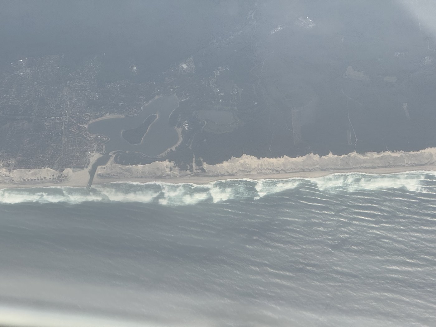 Aus großer Höhe eine Atlantikküste: eine lange Brandungslinie trennt das Meer vom hellen Strand, darüber ziehen dünne Wolken.