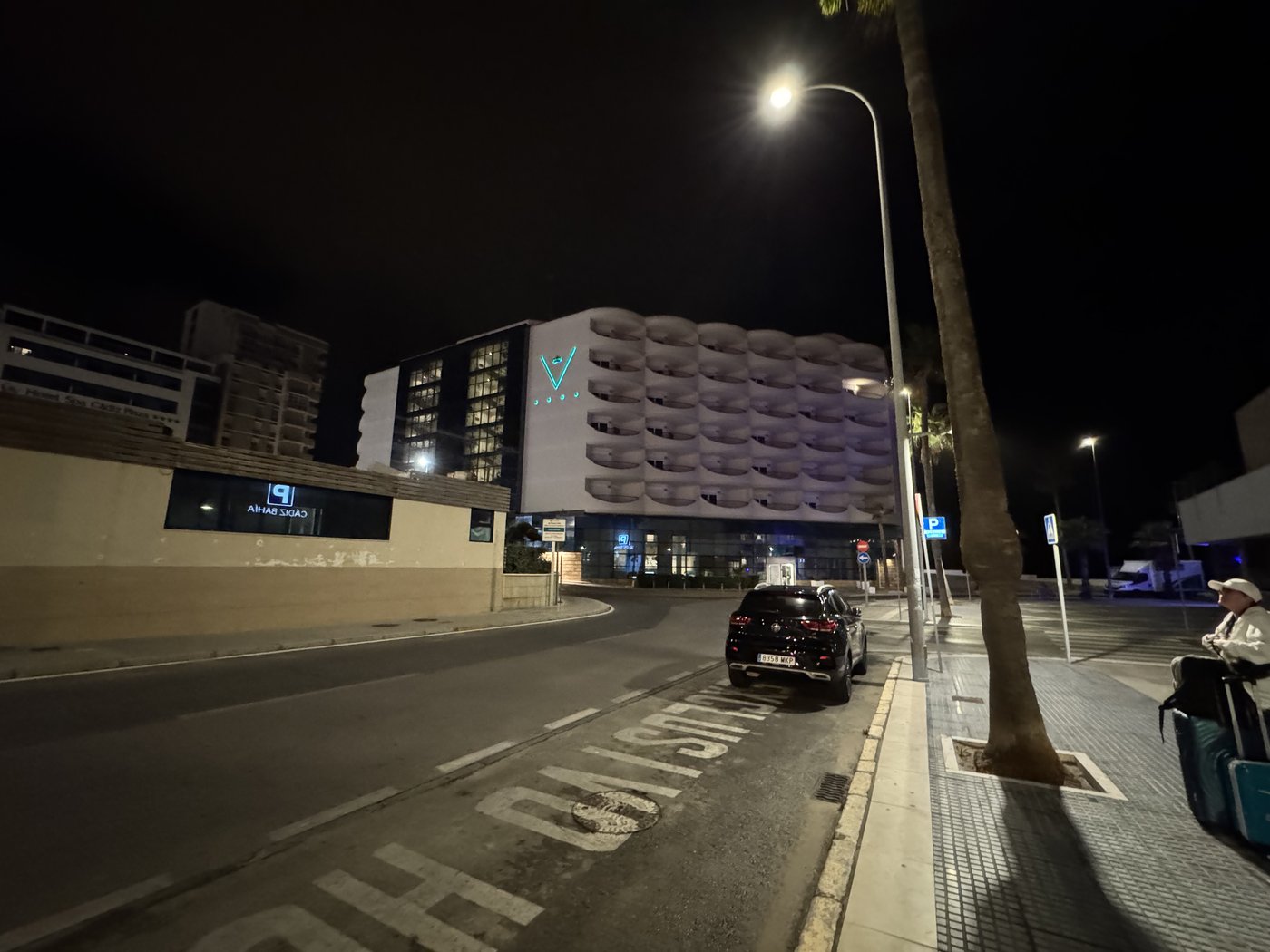 Eine menschenleere Straße vor dem Hotel bei Nacht, nur eine Straßenlaterne und ein geparktes Auto, dahinter beleuchtete Hotelfassaden.