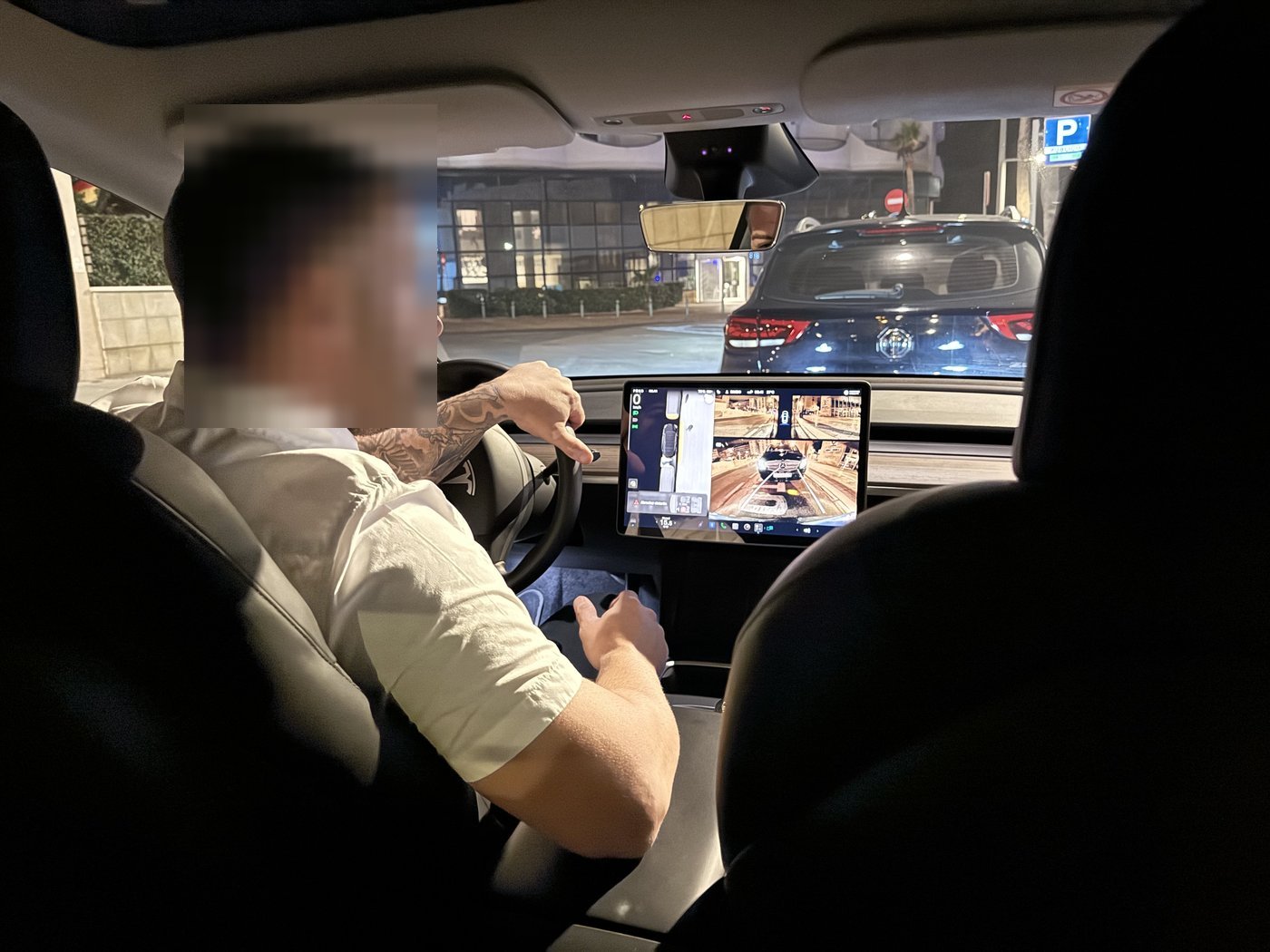 Blick von der Rückbank in den nächtlichen Innenraum des Tesla, der Fahrer am Steuer, der beleuchtete Mittelbildschirm zeigt das Bild der Umgebungskamera.