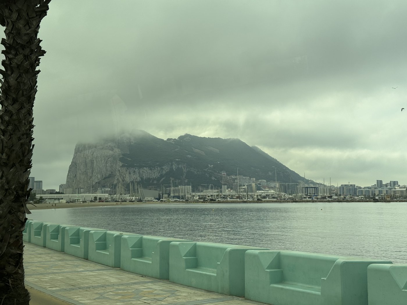 Blick von einer Uferpromenade mit grüner Betonmauer über ruhiges Wasser auf den Felsen von Gibraltar, der unter tiefhängenden Wolken als massiver Rücken aufragt.