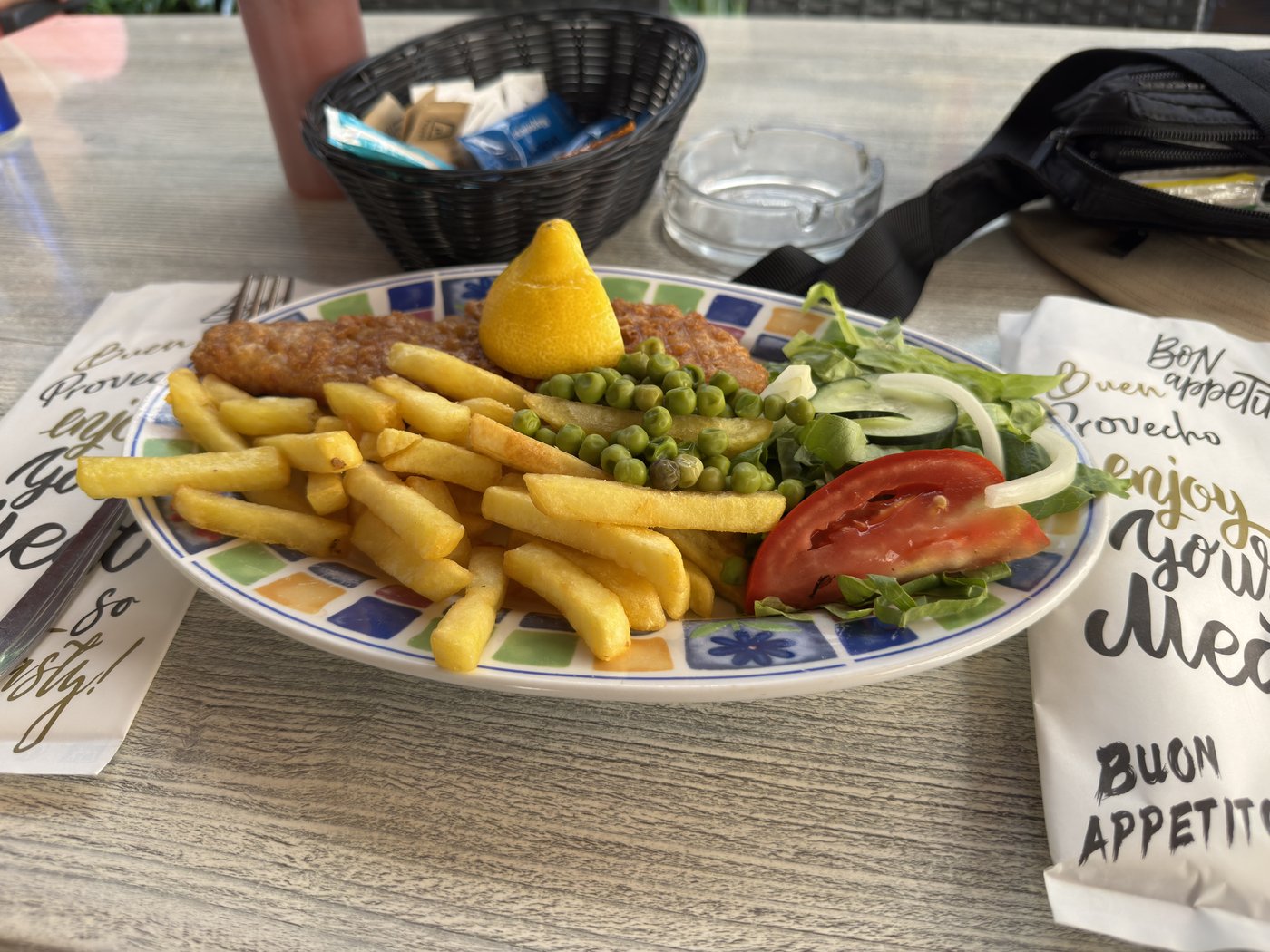 Ein Teller mit Fish and Chips: paniertes Fischfilet, dicke Pommes, ein Zitronenschnitz und Salat, dahinter eine handgeschriebene Tafel.