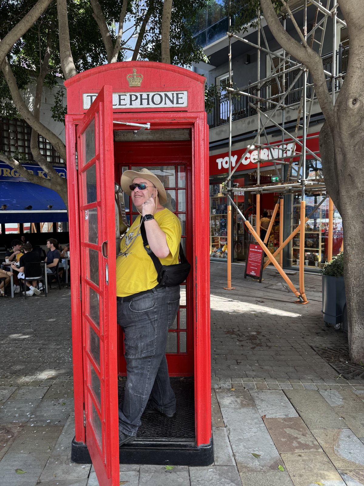 Eine rote britische Telefonzelle am Straßenrand, die Tür geöffnet: ein Mann in gelbem Hemd steht vollständig darin und schaut heraus.