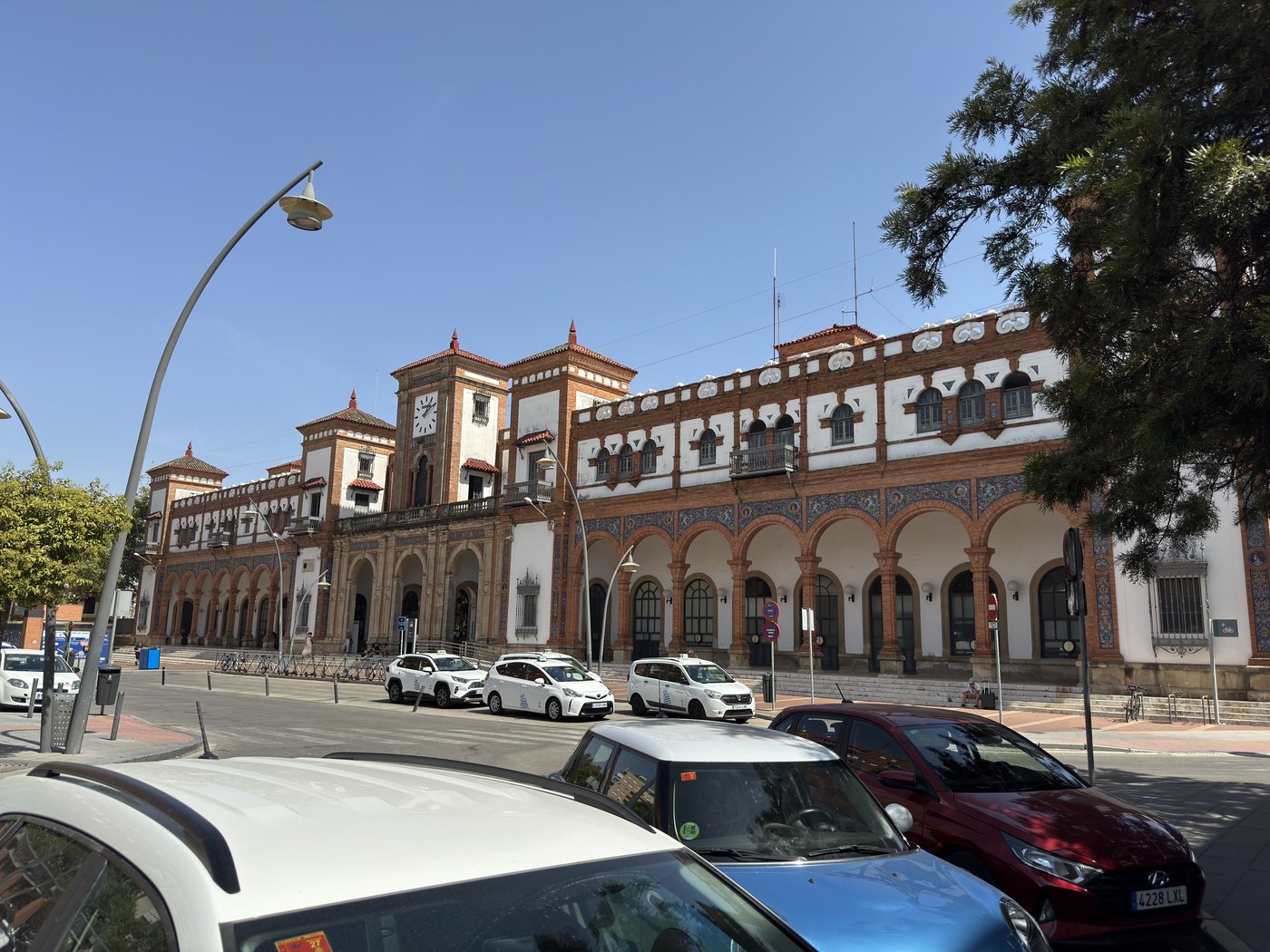 Ein langgestrecktes Bahnhofsgebäude im andalusischen Stil mit Arkadenbögen, Ziegelornamentik und Laternen, davor parkende Autos.