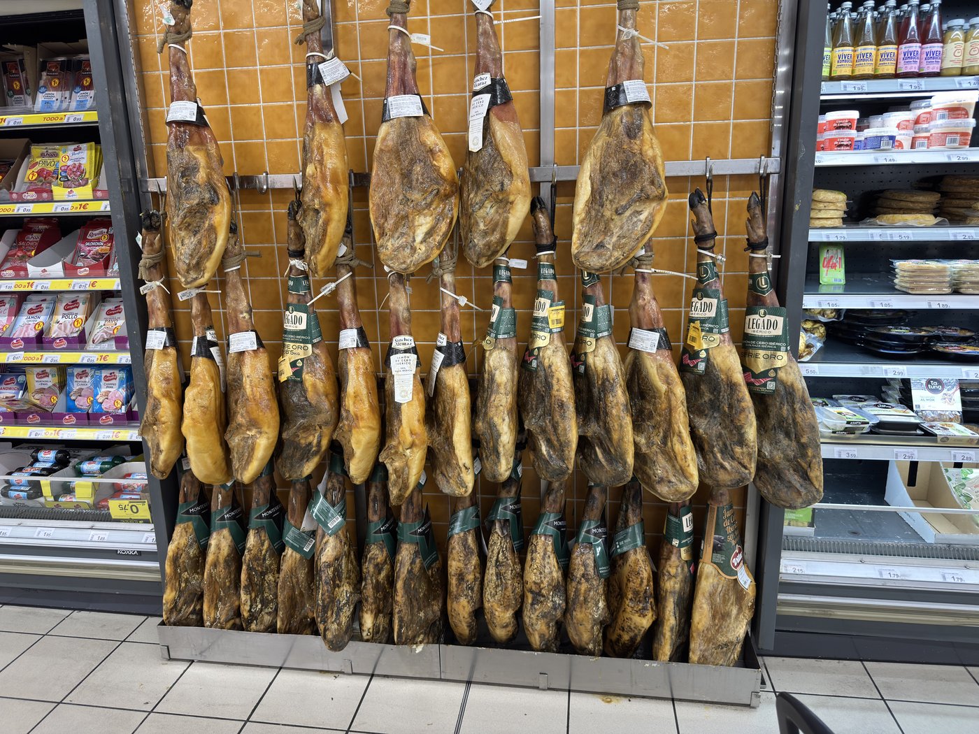 Eine Wand aus Dutzenden ganzen luftgetrockneten Schinken, die dicht an dicht an einer Metallschiene hängen, darunter Supermarktregale.