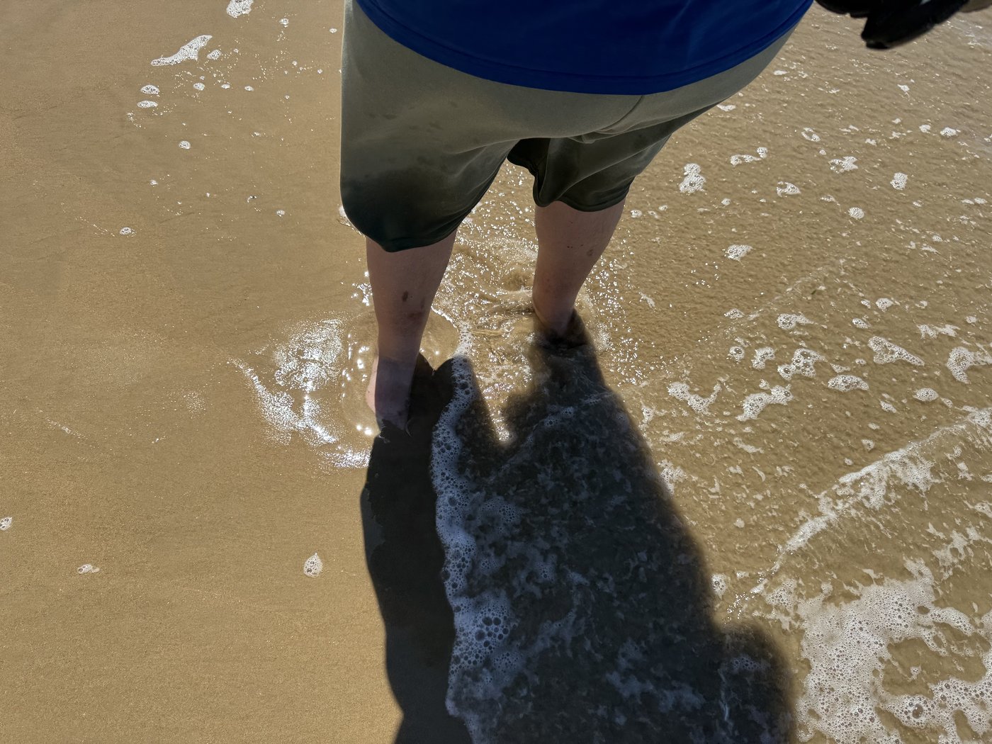 Blick nach unten auf zwei nackte Füße im flachen, auslaufenden Wellenwasser am Sandstrand.
