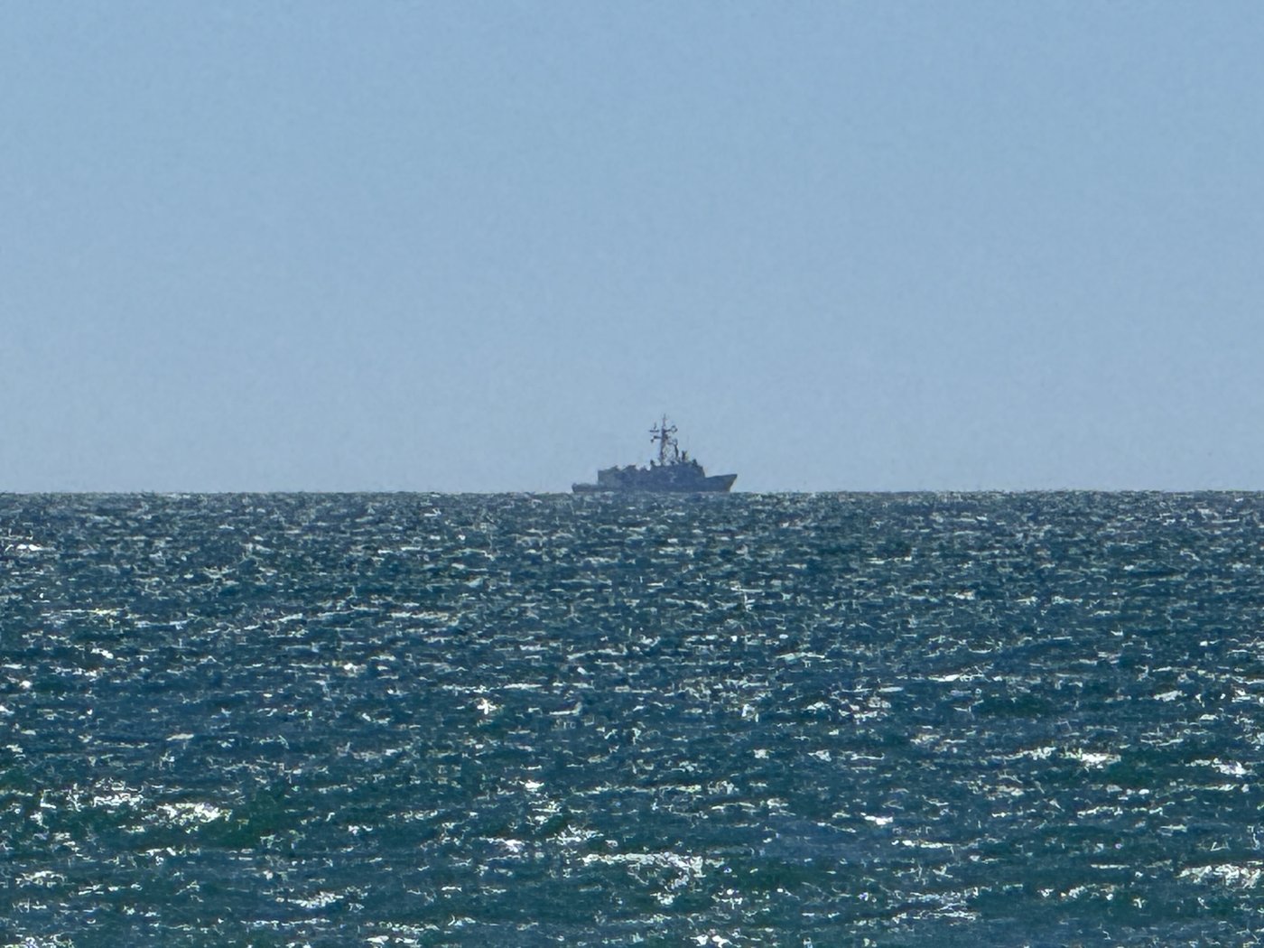 Weit draußen auf dem tiefblauen Meer die graue Silhouette eines Kriegsschiffs vor der scharfen Horizontlinie.