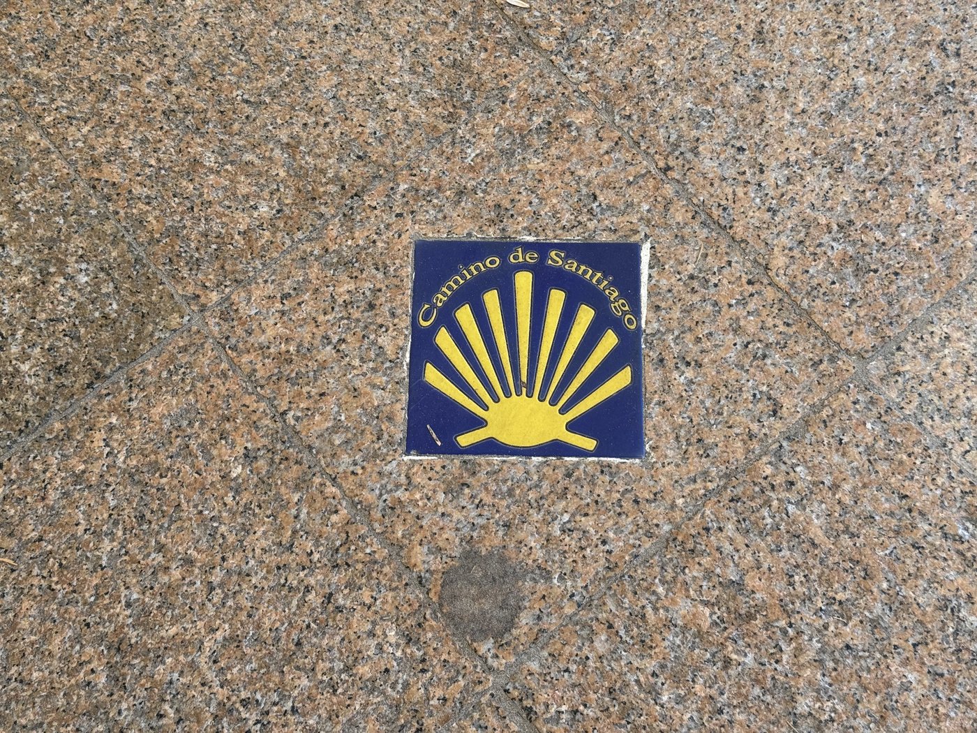 Eine kleine blaue Kachel im rötlichen Granitpflaster des Gehwegs, darauf die gelbe stilisierte Jakobsmuschel und die Aufschrift Camino de Santiago.