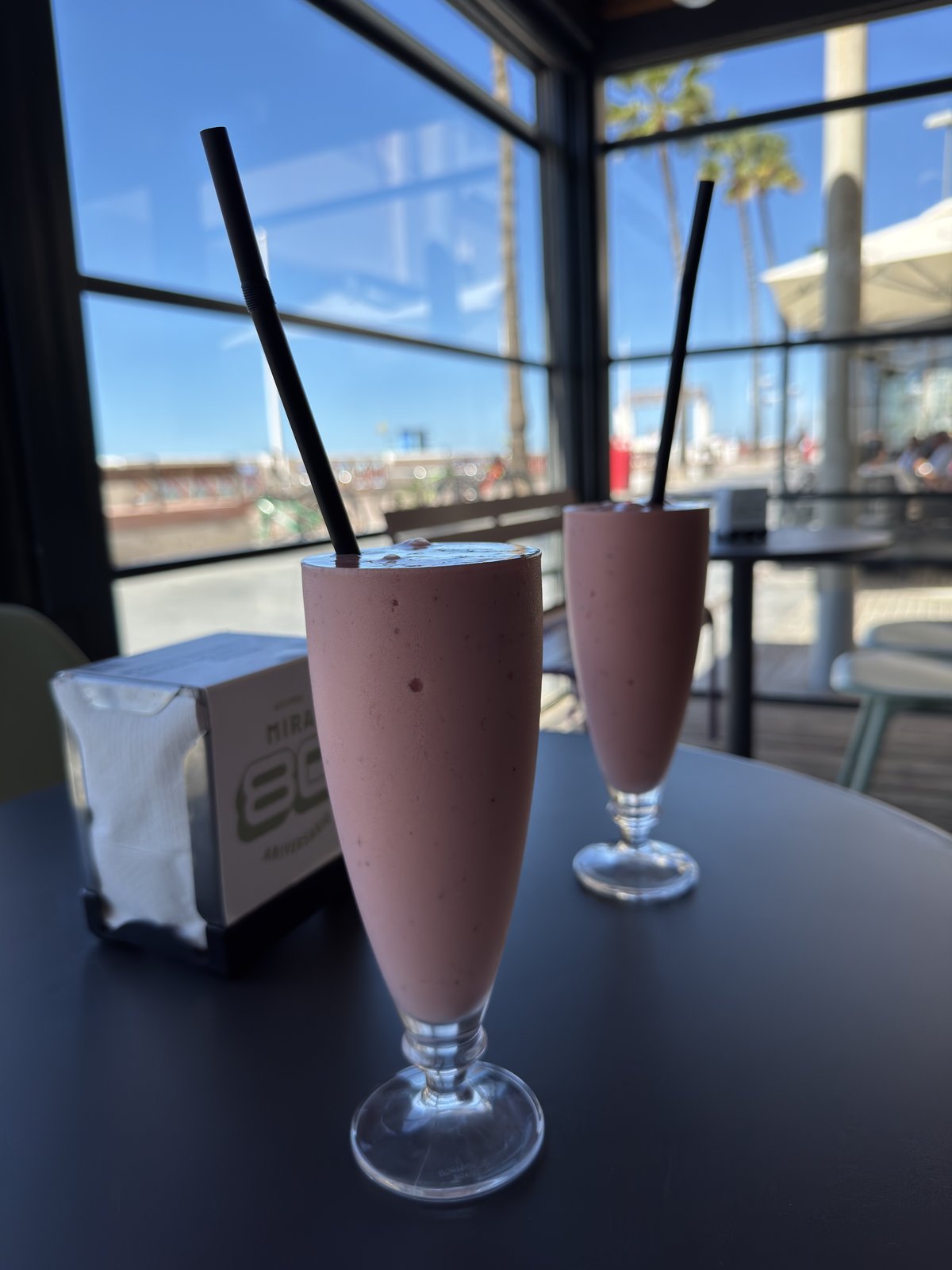 Zwei hohe Gläser mit rosafarbenem Erdbeermilchshake und schwarzen Trinkhalmen auf einem dunklen Tisch, dahinter eine Fensterfront mit Palmen und Meer.