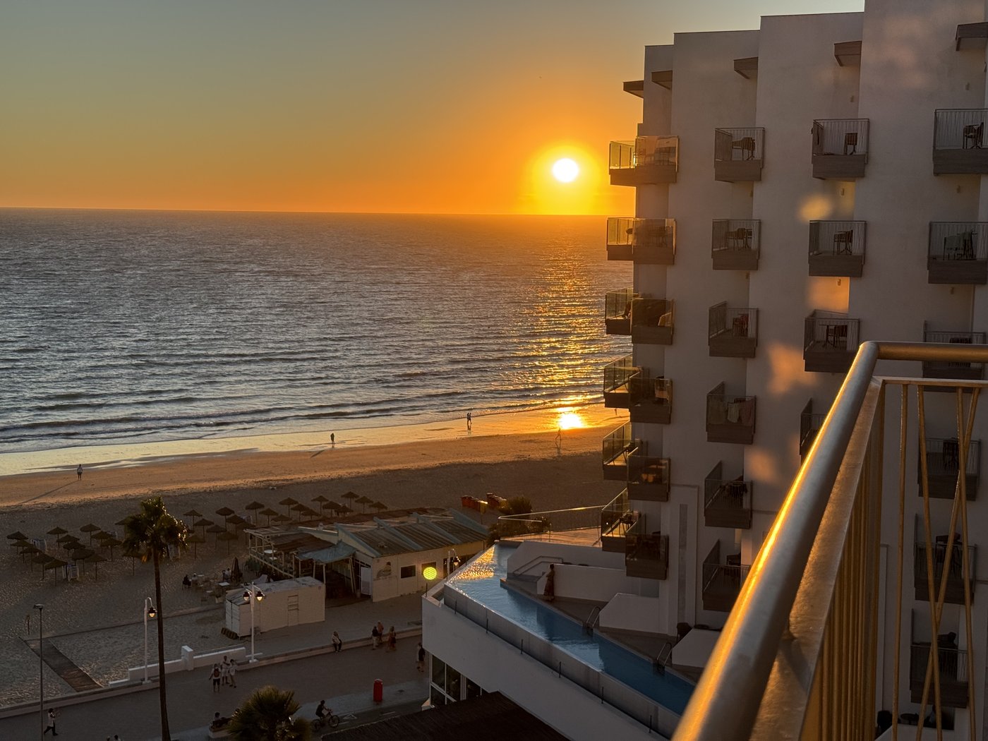 Die untergehende Sonne als gleißender Ball dicht über dem Meer, der Himmel in Orange und Gold, rechts im Bild die Silhouette des Nachbarhotels.