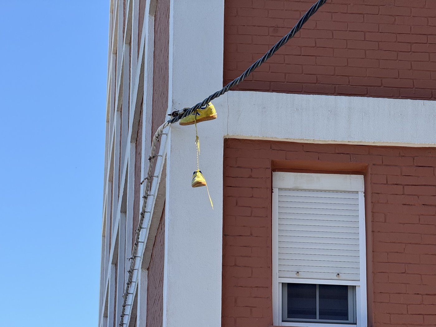 An einem Kabel vor einer weiß-roten Hausfassade hängt ein Paar gelber Schuhe, zusammengeknotet an den Schnürsenkeln, vor blauem Himmel.