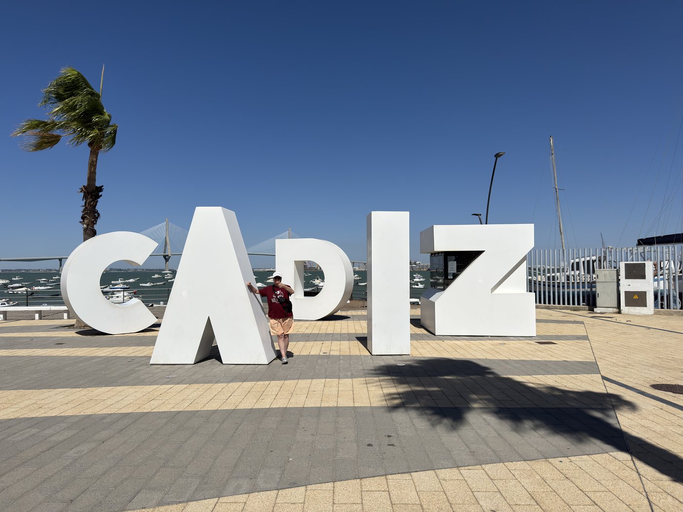 Die überlebensgroßen weißen Buchstaben des Schriftzugs CÁDIZ auf der gepflasterten Promenade, dahinter eine Palme im Wind.
