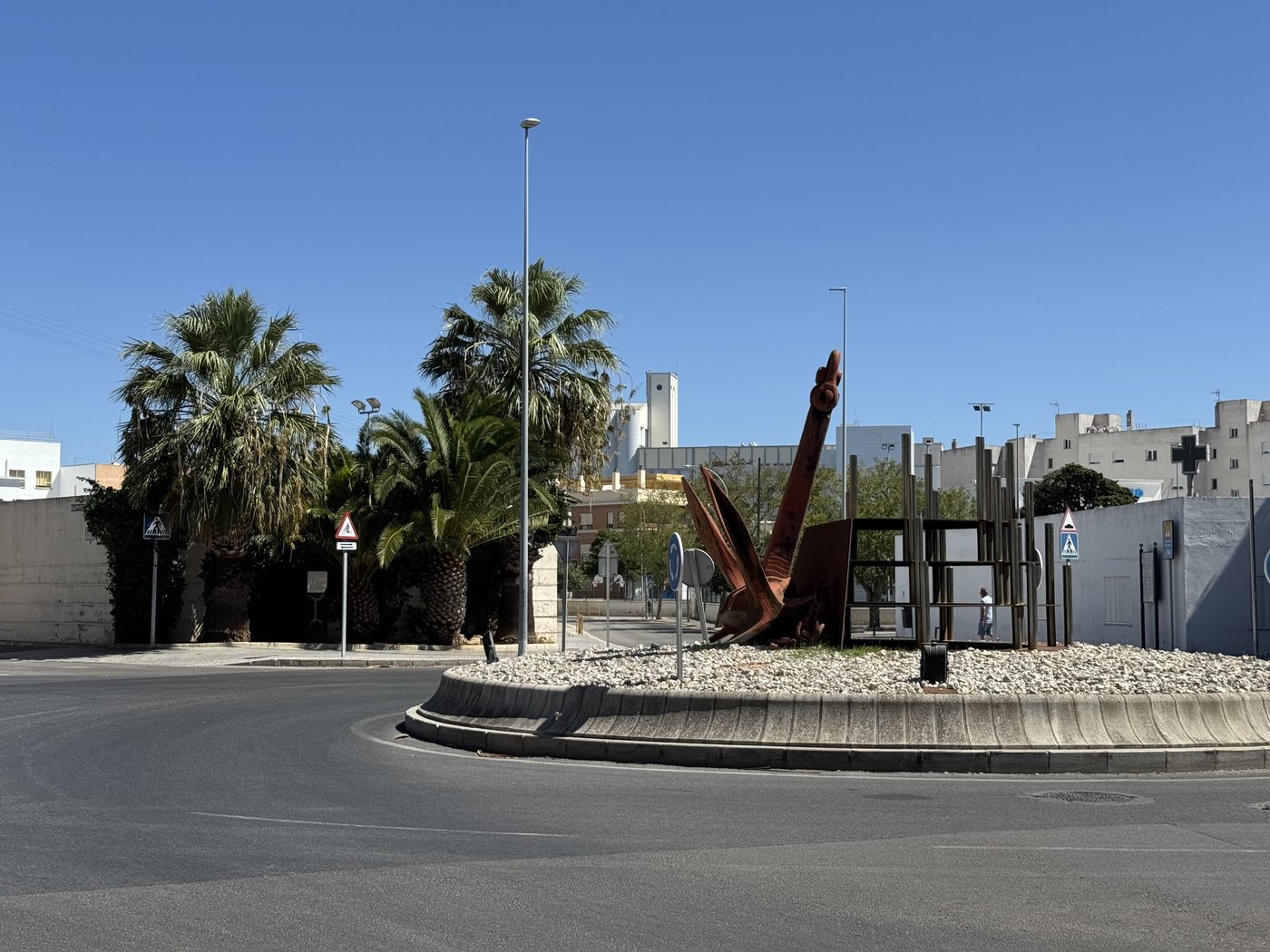 Auf einer Verkehrsinsel steht ein großer rostroter Patentanker als Denkmal, dahinter Palmen und helle Gebäude.