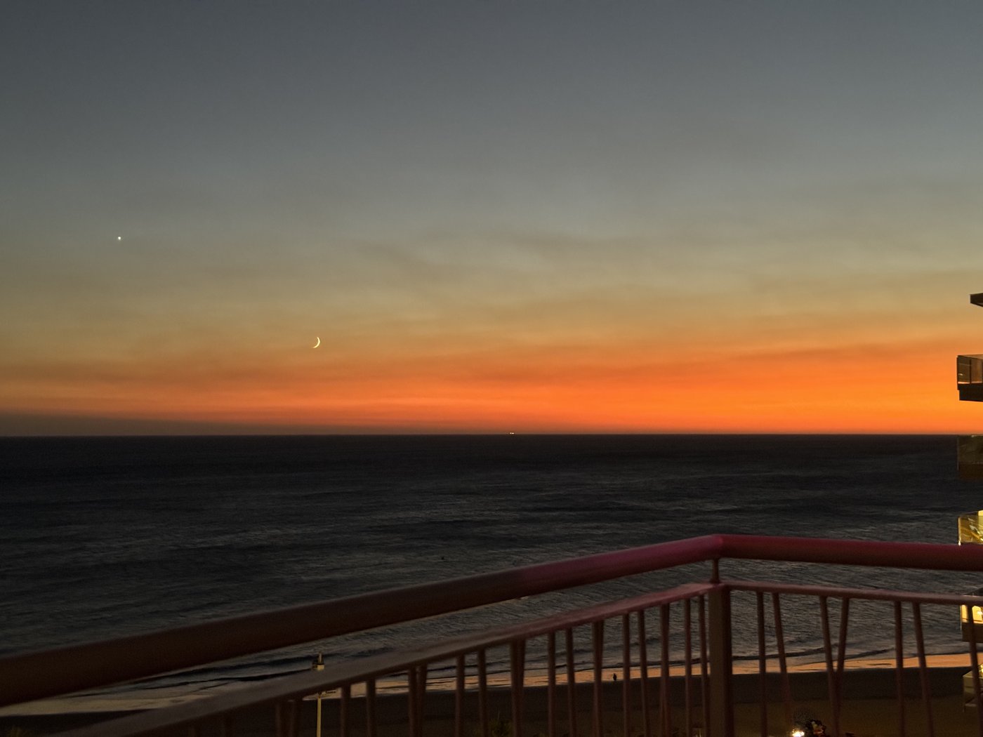 Sonnenuntergang über dem Meer: ein breites orangefarbenes Band am Horizont unter dunkelblauem Himmel, im Vordergrund die Silhouette des Balkongeländers.