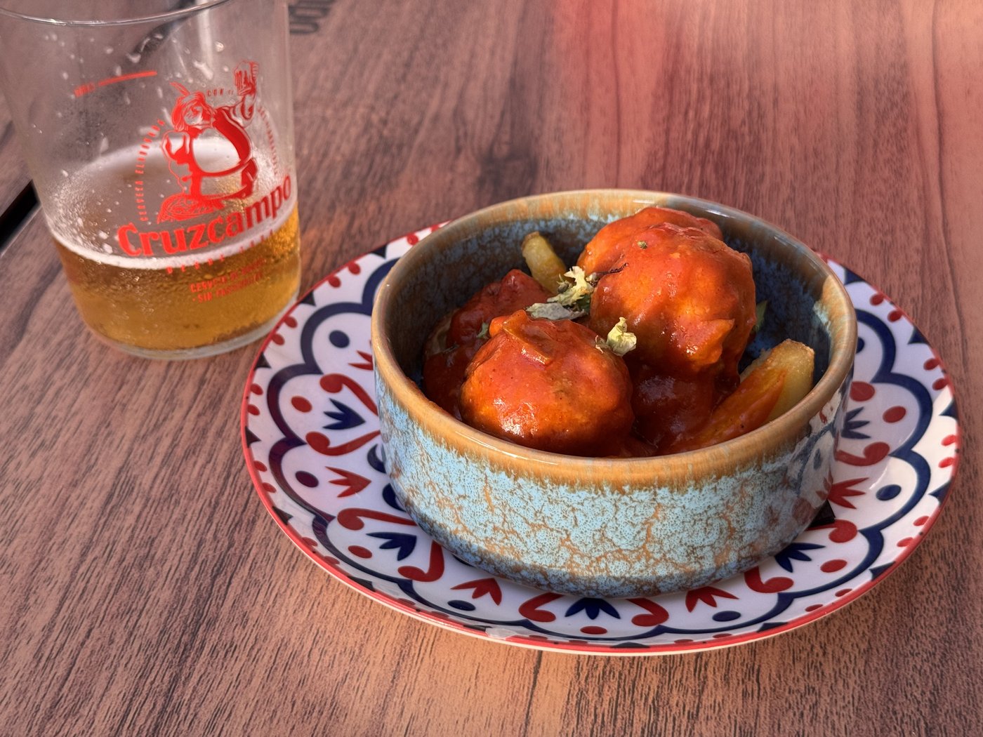 Eine türkis gemusterte Schale mit Thunfischbällchen in dunkler Tomatensauce auf einem rot-blau gemusterten Teller, daneben ein halbvolles Bierglas auf einem Holztisch.