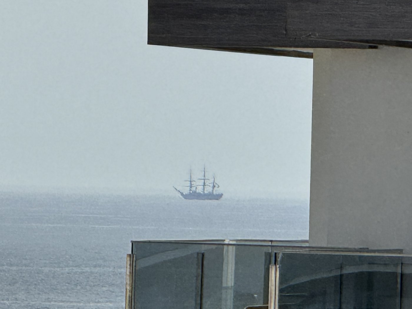 Blick zwischen Balkonbrüstung und Deckenkante hindurch aufs diesige Meer: weit draußen die kleine Silhouette eines Großseglers mit drei Masten.