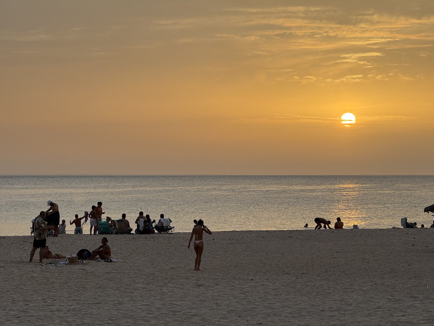Die Sonne steht tief über dem Meer und färbt Himmel und Wasser orange; am Strand sitzen einzelne Menschen im Gegenlicht.