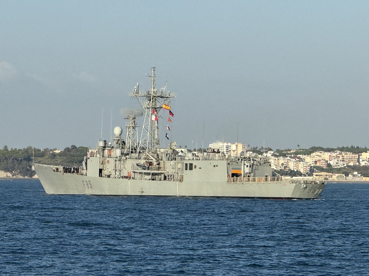 Eine graue Fregatte der spanischen Marine läuft bei ruhiger See vor einer flachen Küstenlinie ein.