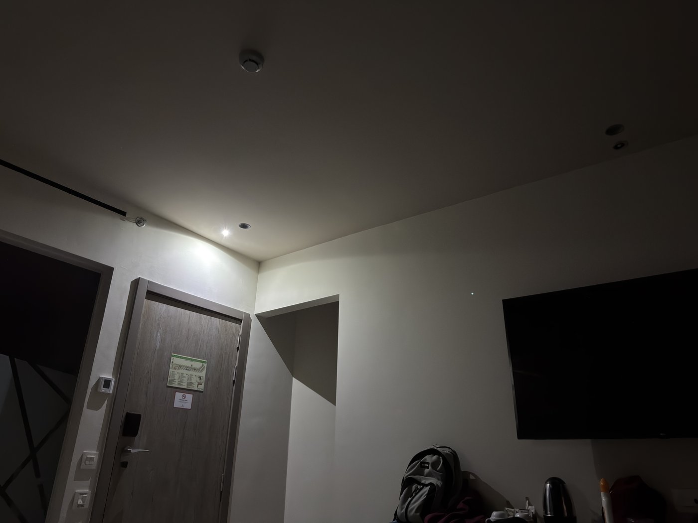 Ein abgedunkeltes Hotelzimmer, in dem über der Tür eine einzelne helle Leuchte brennt und den Raum ungleichmäßig ausleuchtet.