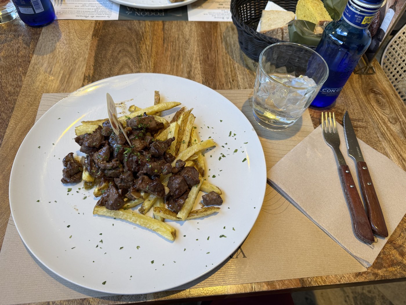 Ein Teller mit grob geschnittenen Rindfleischstücken auf Pommes frites, daneben Besteck und Gläser auf einer Holztischplatte.