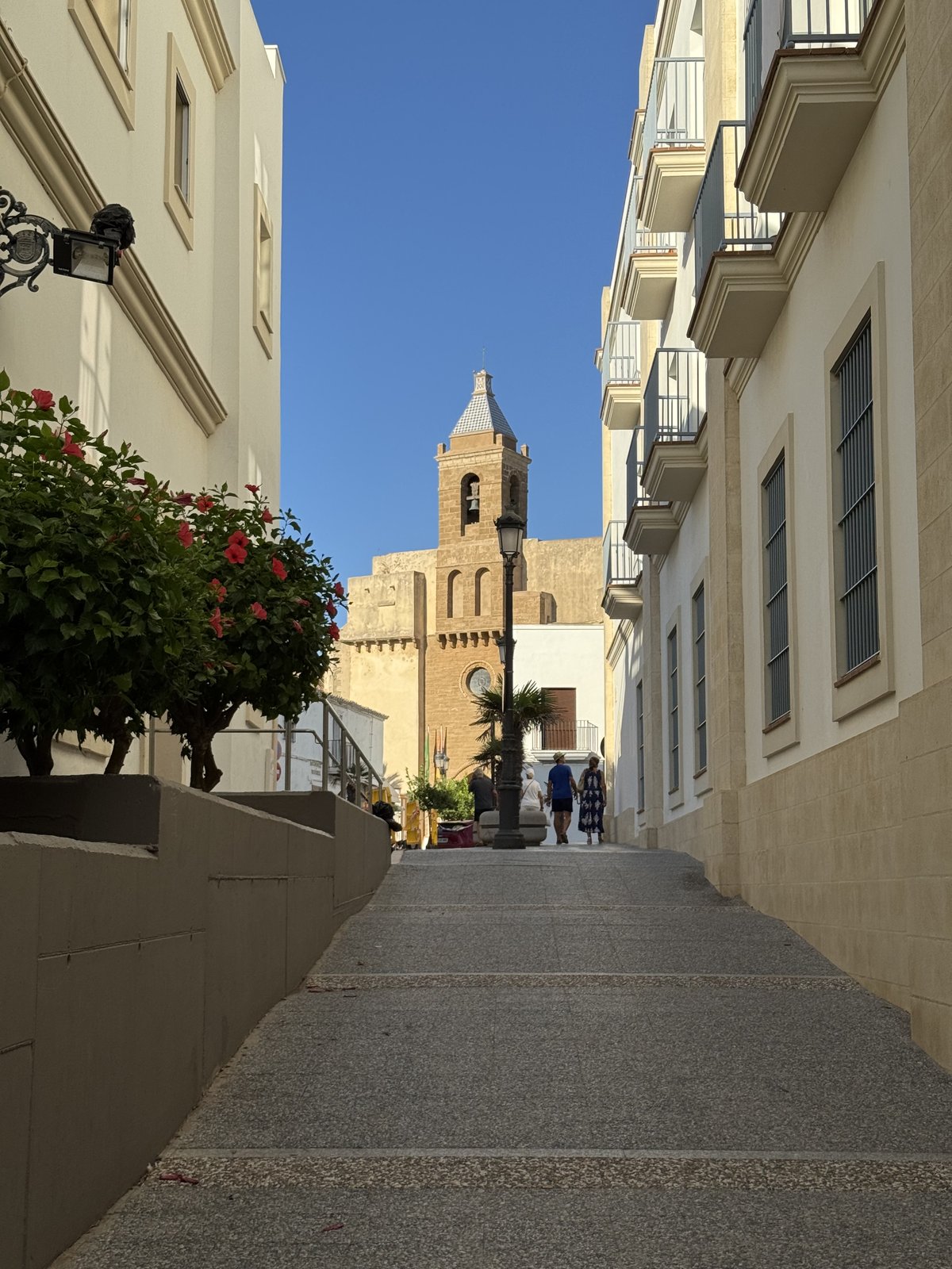 Eine schmale, gepflasterte Gasse zwischen weißen Häusern, am Ende ein heller Kirchturm vor blauem Himmel.