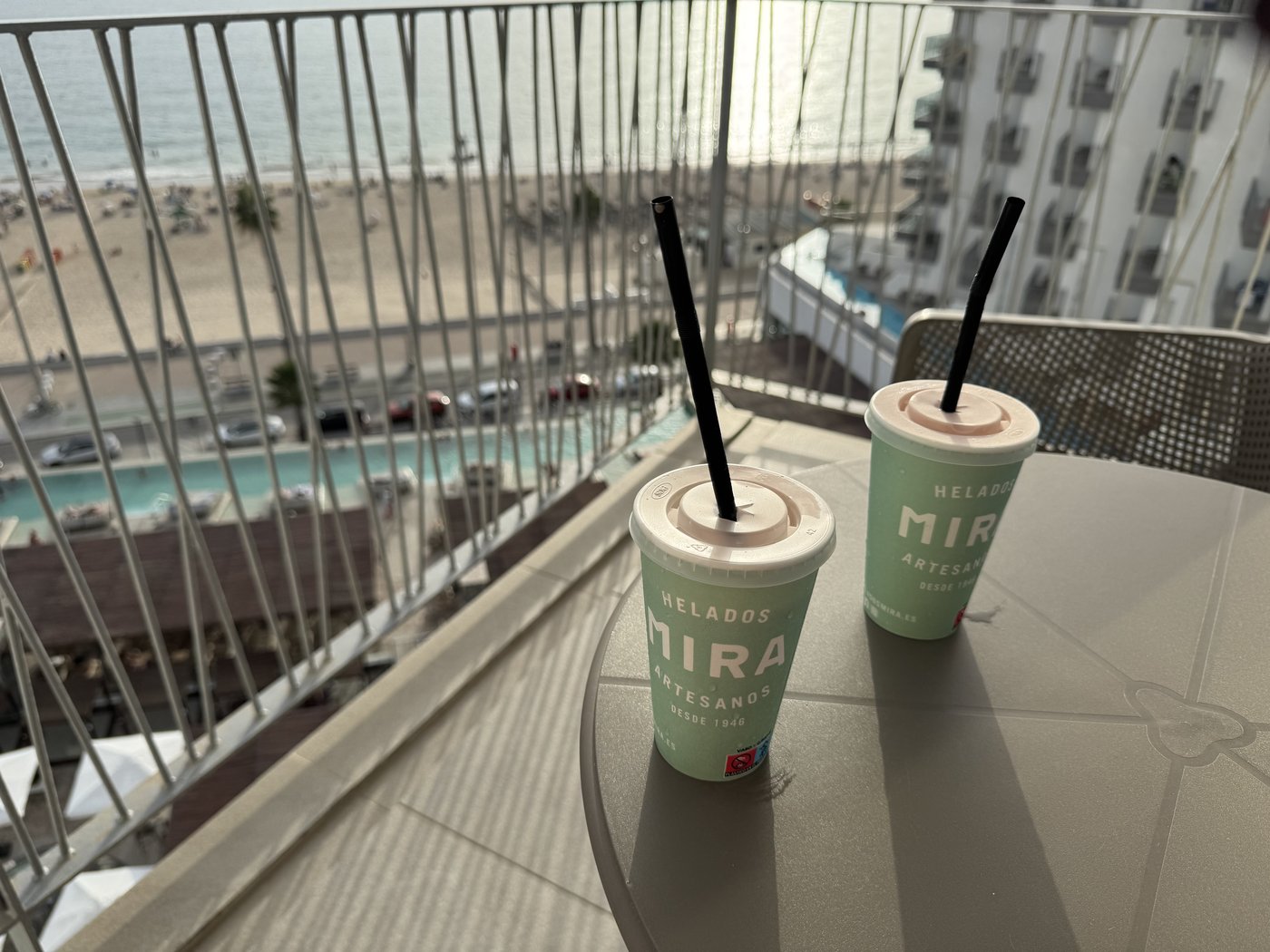Zwei Becher mit hellem Milchshake und Trinkhalmen stehen auf einem Balkongeländer, dahinter der Pool und das Meer.