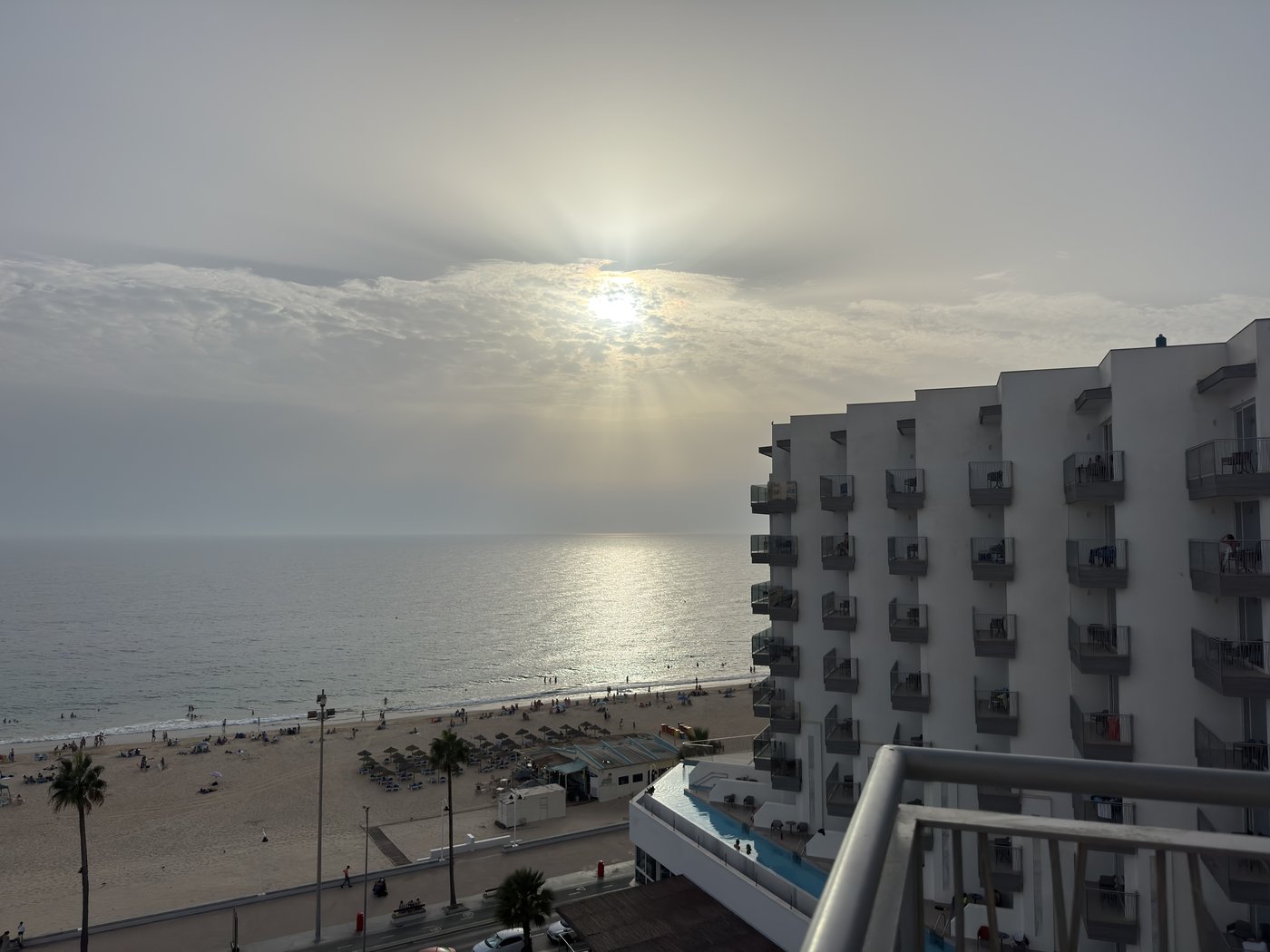 Ein bewölkter Sonnenuntergang über dem Meer, die Sonne steht als heller Fleck hinter einer Wolkendecke über dem Strand.