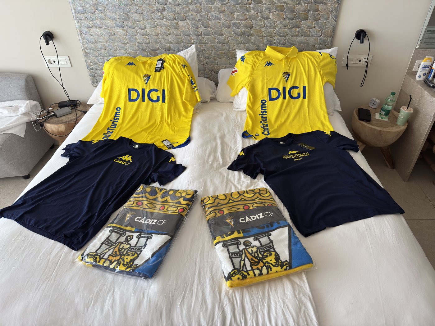 Auf einem Hotelbett ausgebreitet: zwei gelbe Trikots, zwei dunkelblaue T-Shirts und mehrere gelb-blaue Vereinsschals des Cádiz CF.