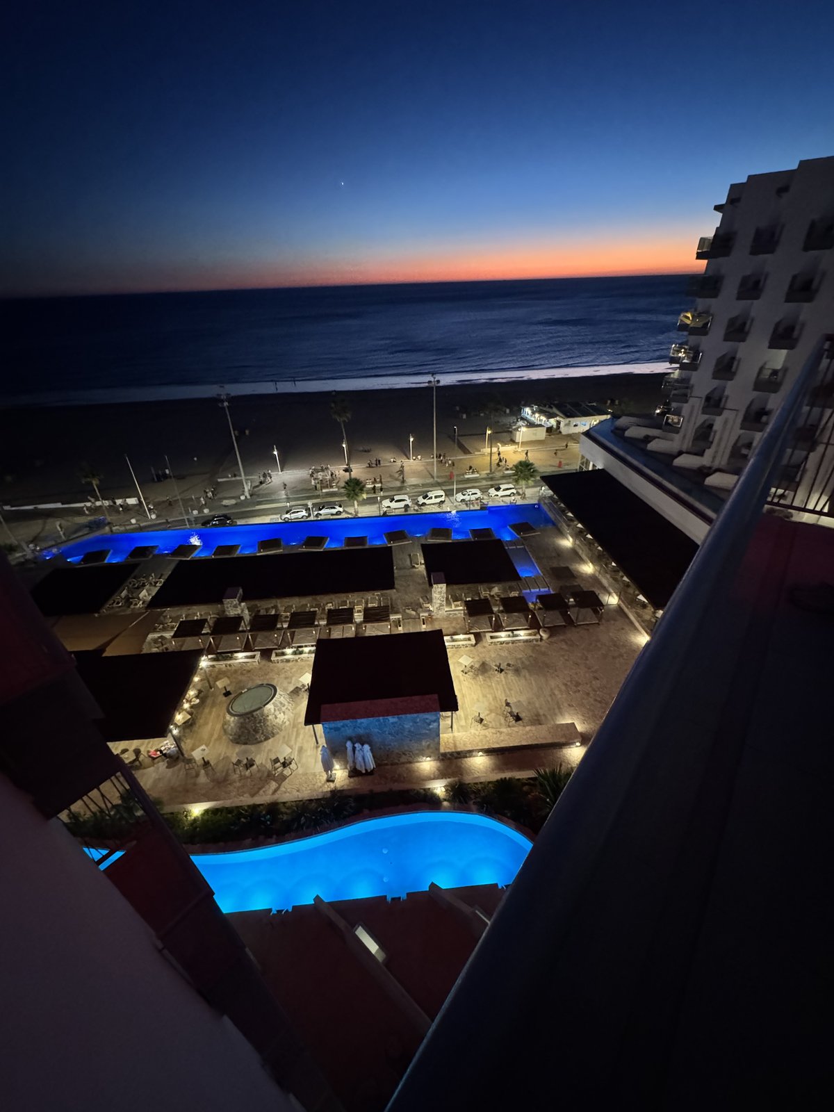 Blick vom Balkon zur blauen Stunde: der beleuchtete Hotelpool leuchtet türkis aus der Dunkelheit, dahinter das Meer und ein letzter oranger Streifen am Horizont.