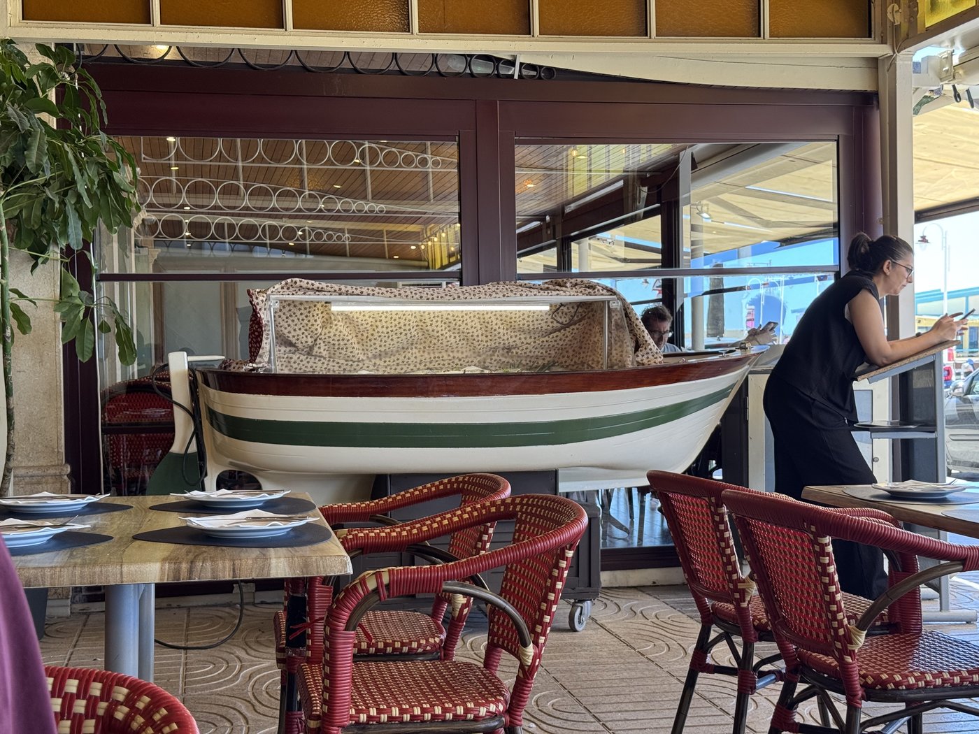 Im Gastraum steht ein kleines hölzernes Ruderboot als Auslage für frischen Fisch, davor Tische mit roten Korbstühlen, im Hintergrund die offene Front zum Strand.