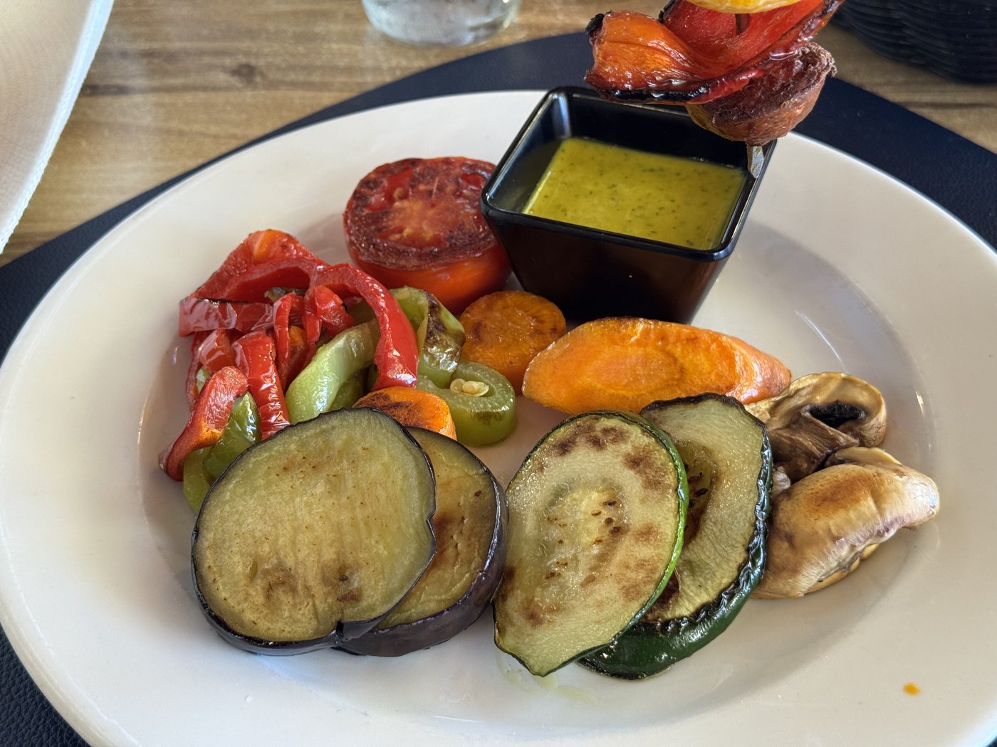 Ein weißer Teller mit gegrillten Auberginen- und Zucchinischeiben, Paprika, Karotte und Tomate, dazu ein schwarzes Schälchen mit heller Soße.