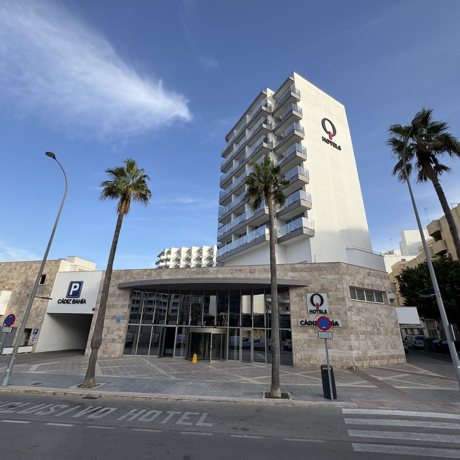 Ein weißes Hotelhochhaus hinter hohen Palmen an einer leeren Straßenkreuzung im Morgenlicht.