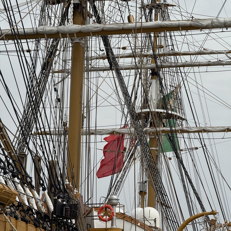 Blick senkrecht in die Takelage eines Großseglers: Masten, Rahen und ein dichtes Netz aus Tauwerk, dazwischen eine rote Flagge.