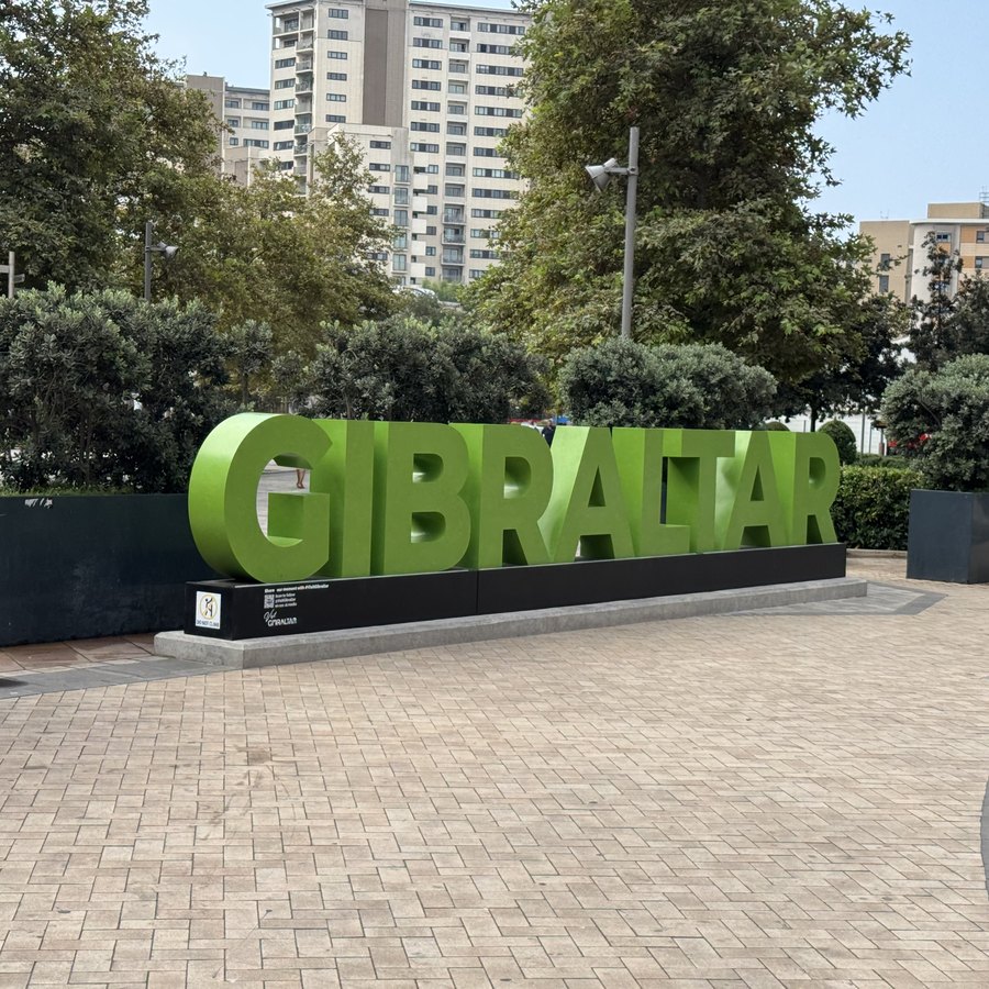 Große begrünte Buchstaben bilden den Schriftzug GIBRALTAR auf einem gepflasterten Platz, dahinter Hochhäuser.