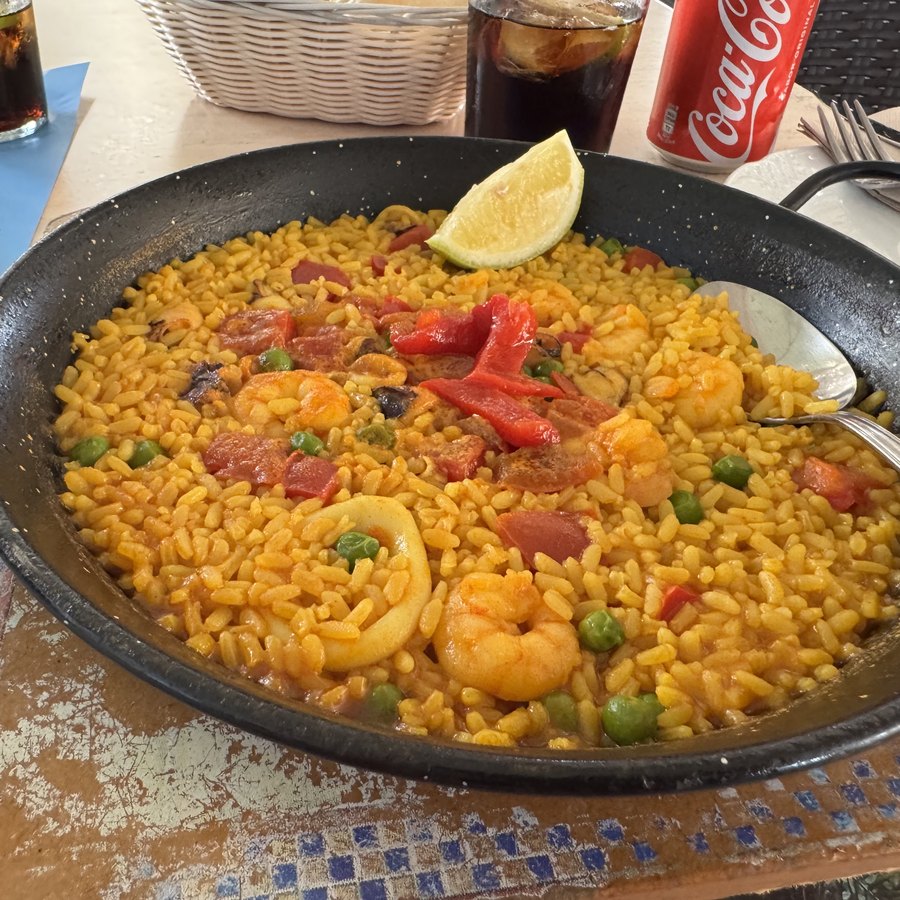 Eine Pfanne Paella mit Garnelen, Muscheln und Zitrone, daneben ein Glas und eine Cola-Dose.